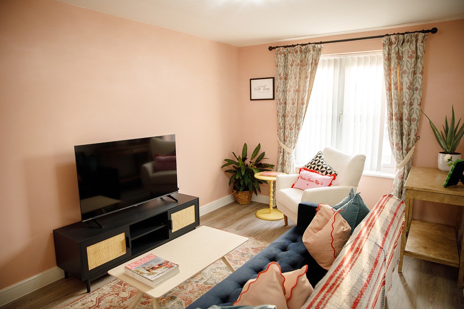 A cozy living room with a peach-colored wall, a flat-screen TV on a black entertainment center, a white armchair with decorative pillows, a dark gray sofa with pillows, a window with floral curtains, a small yellow side table, and several potted plan