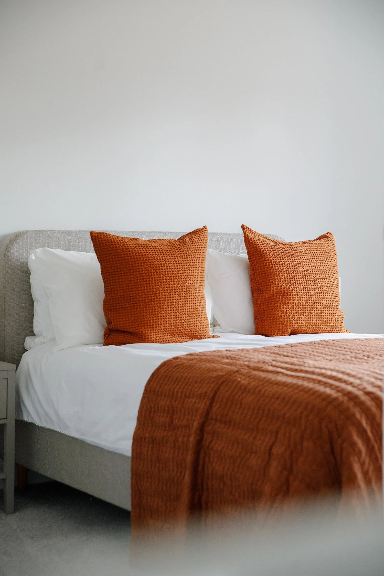 A neatly made bed with white sheets and pillows, two orange textured throw pillows, and an orange bedspread, against a plain white wall.