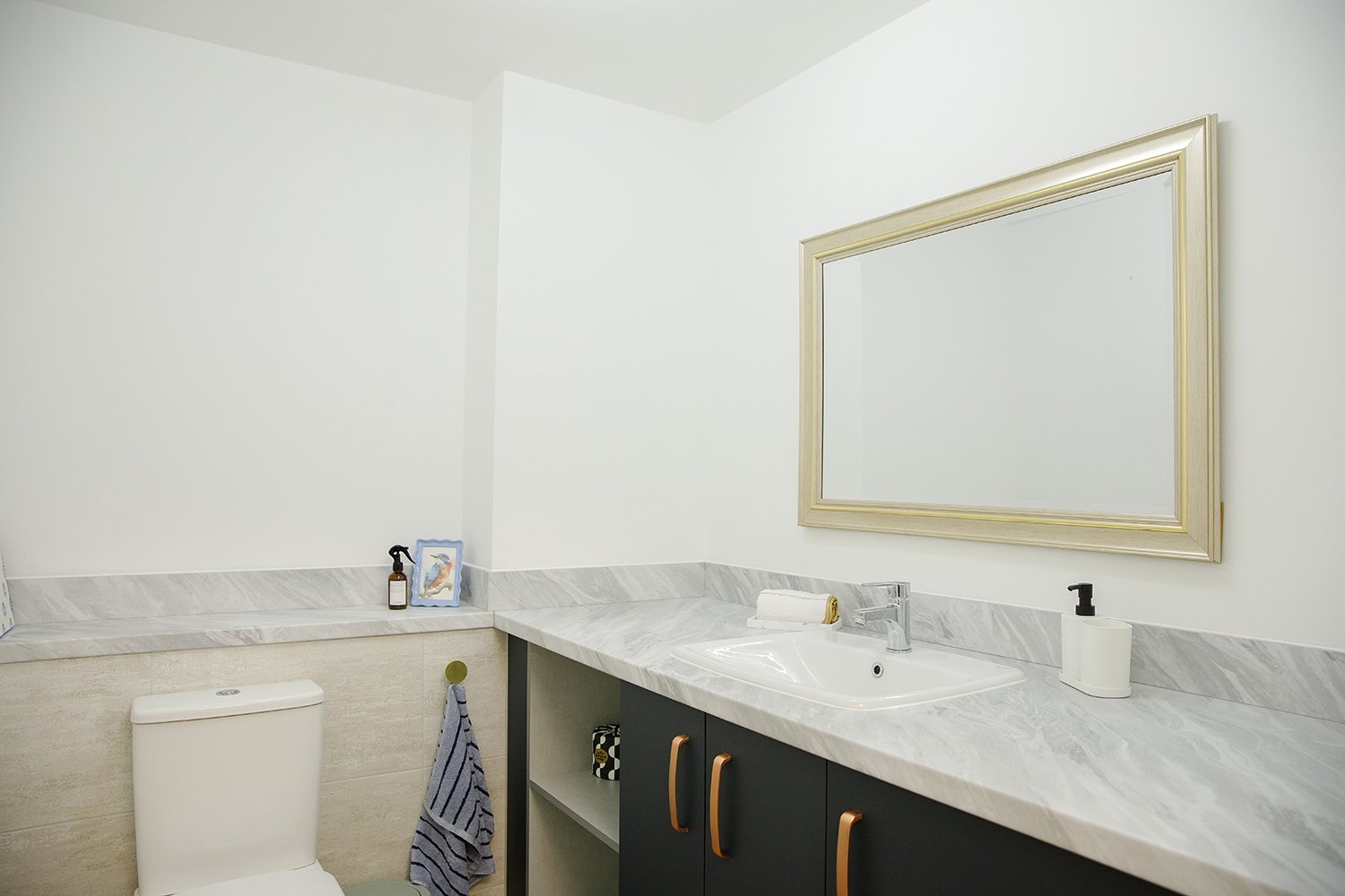 Bathroom with white walls, marble countertop, large mirror, black cabinet with gold handles, white toilet, towel, soap dispenser, bottle, and framed picture on the countertop.