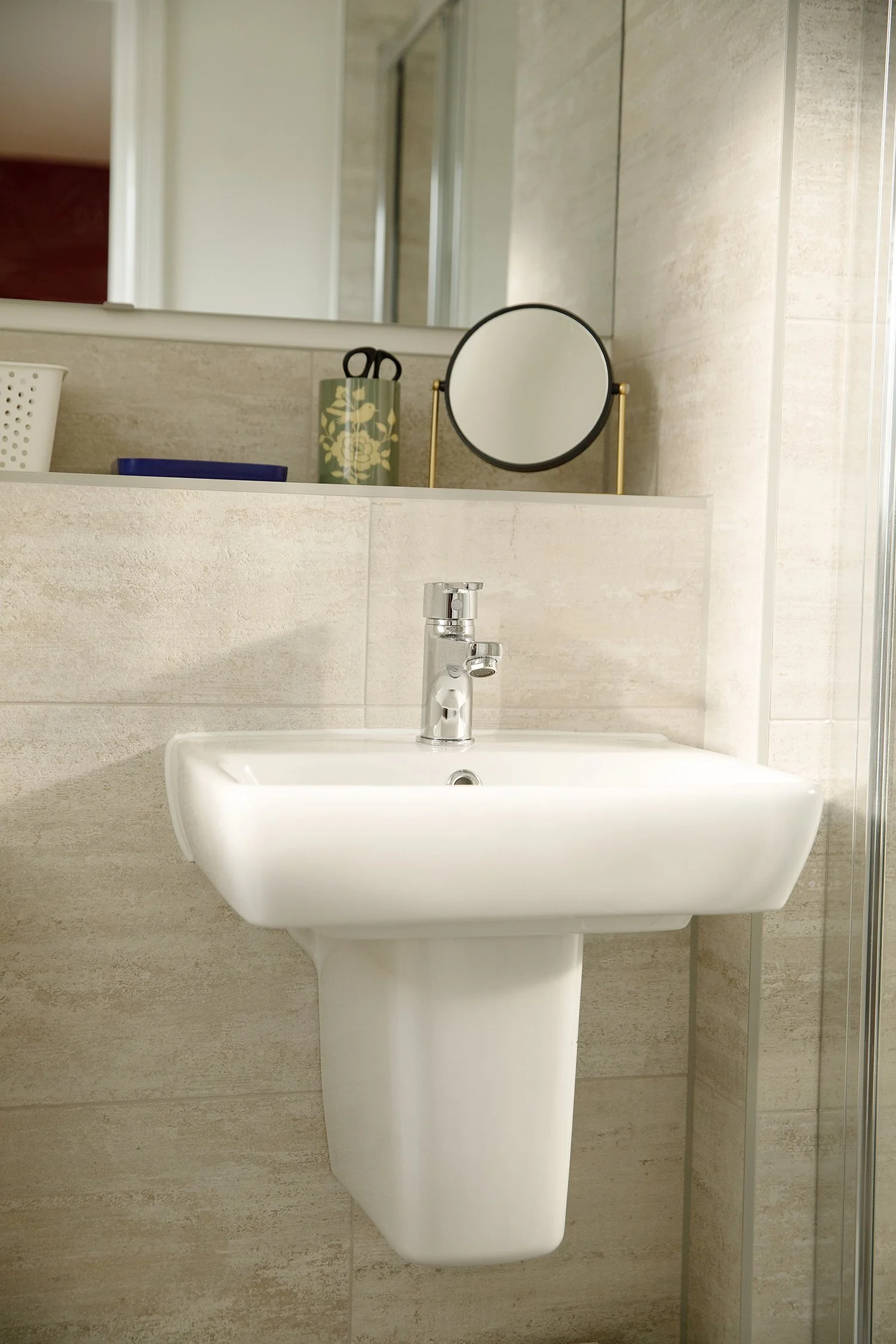 A bathroom sink with a silver faucet, mounted on a beige tiled wall. Above the sink, a large mirror with part of the reflection visible, showing a door and other parts of the bathroom. To the right of the mirror, on the countertop, there is a small r