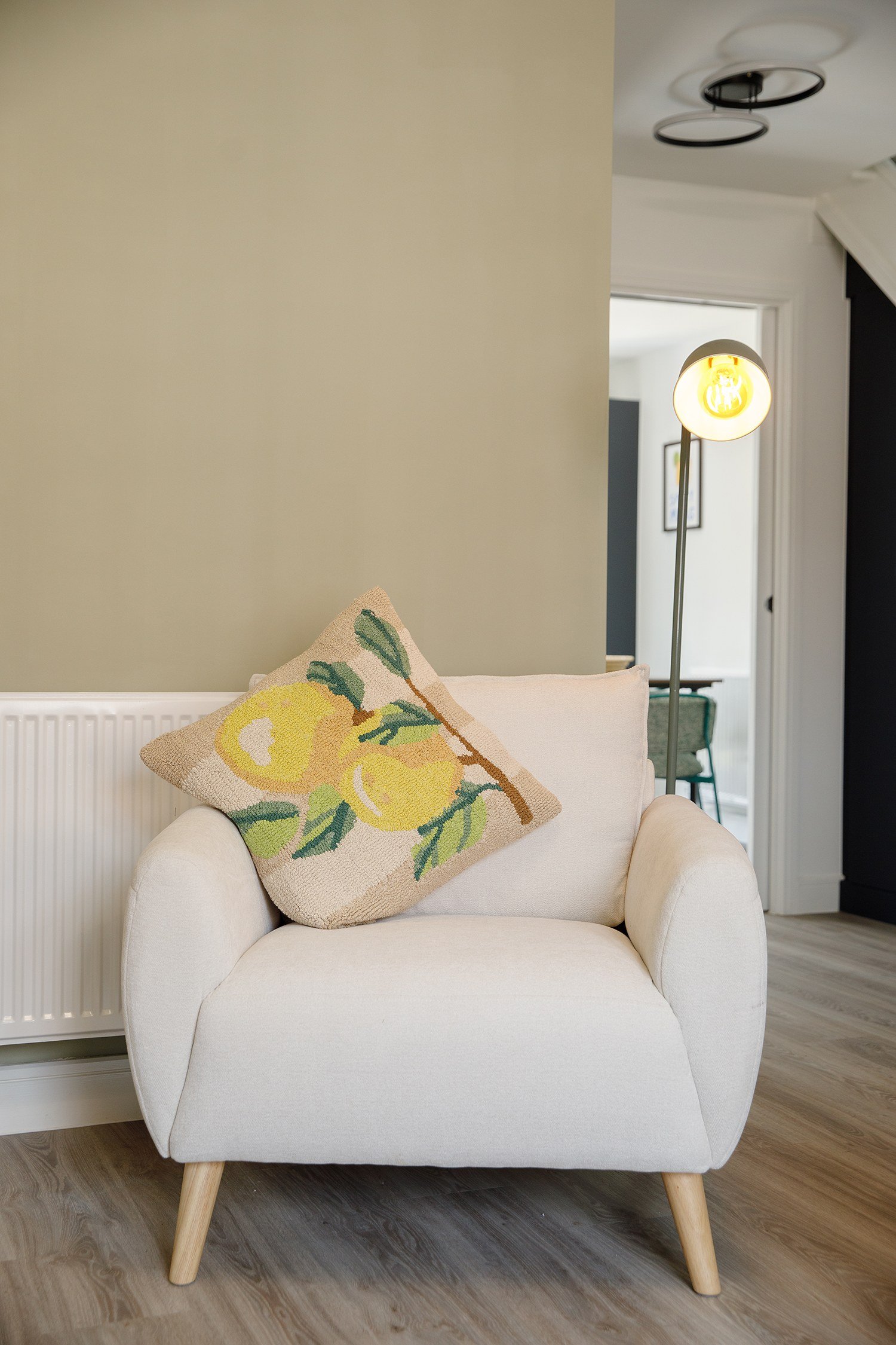A white armchair with rounded arms and wooden legs, placed on a wooden floor. A decorative pillow with a yellow pear and green leaves pattern is on the chair. A standing lamp with a round, warm light bulb is next to it. In the background, there is a 