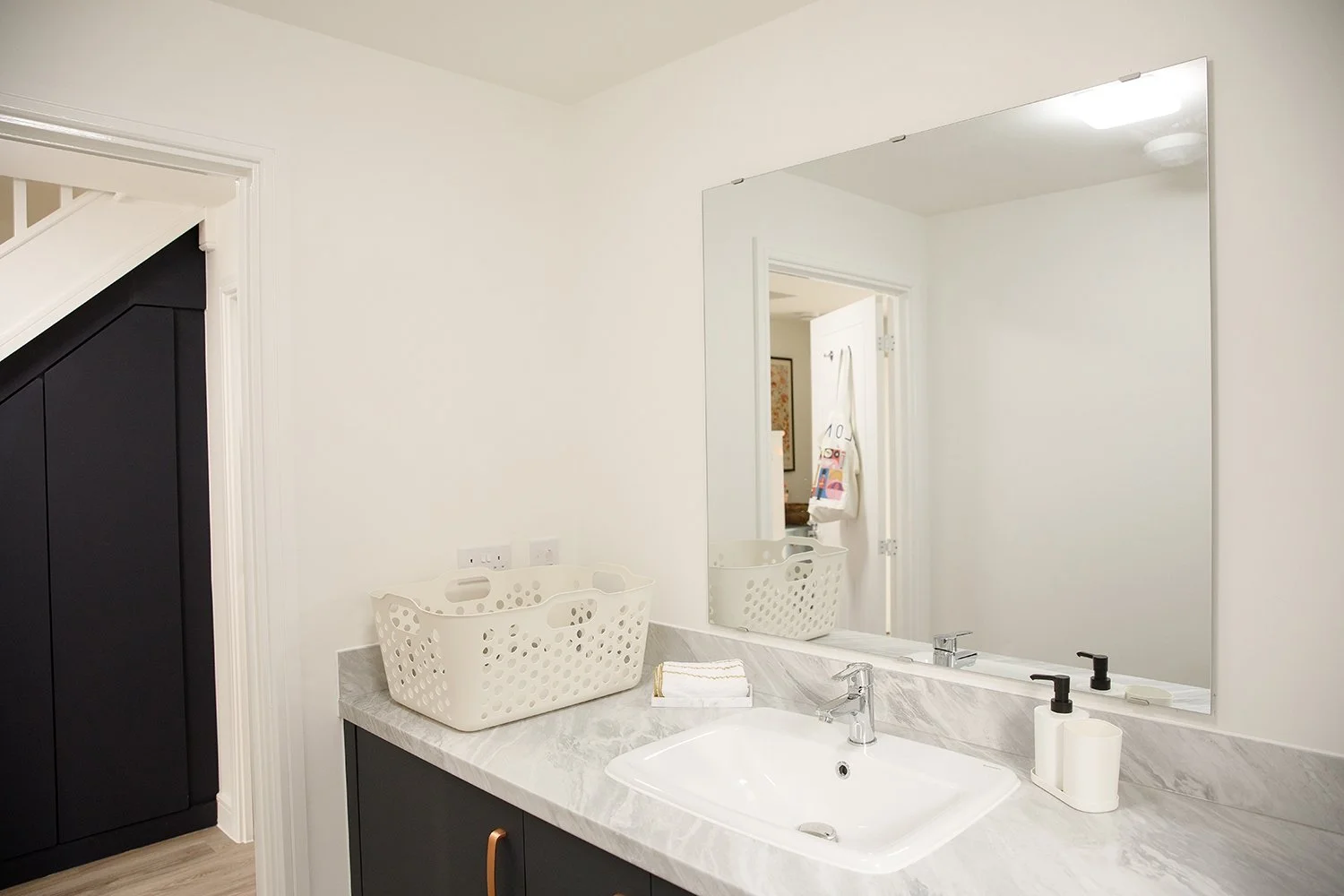 A bathroom with a white countertop, a large mirror, a white sink, a soap dispenser, folded towels, a laundry basket, and black cabinetry.
