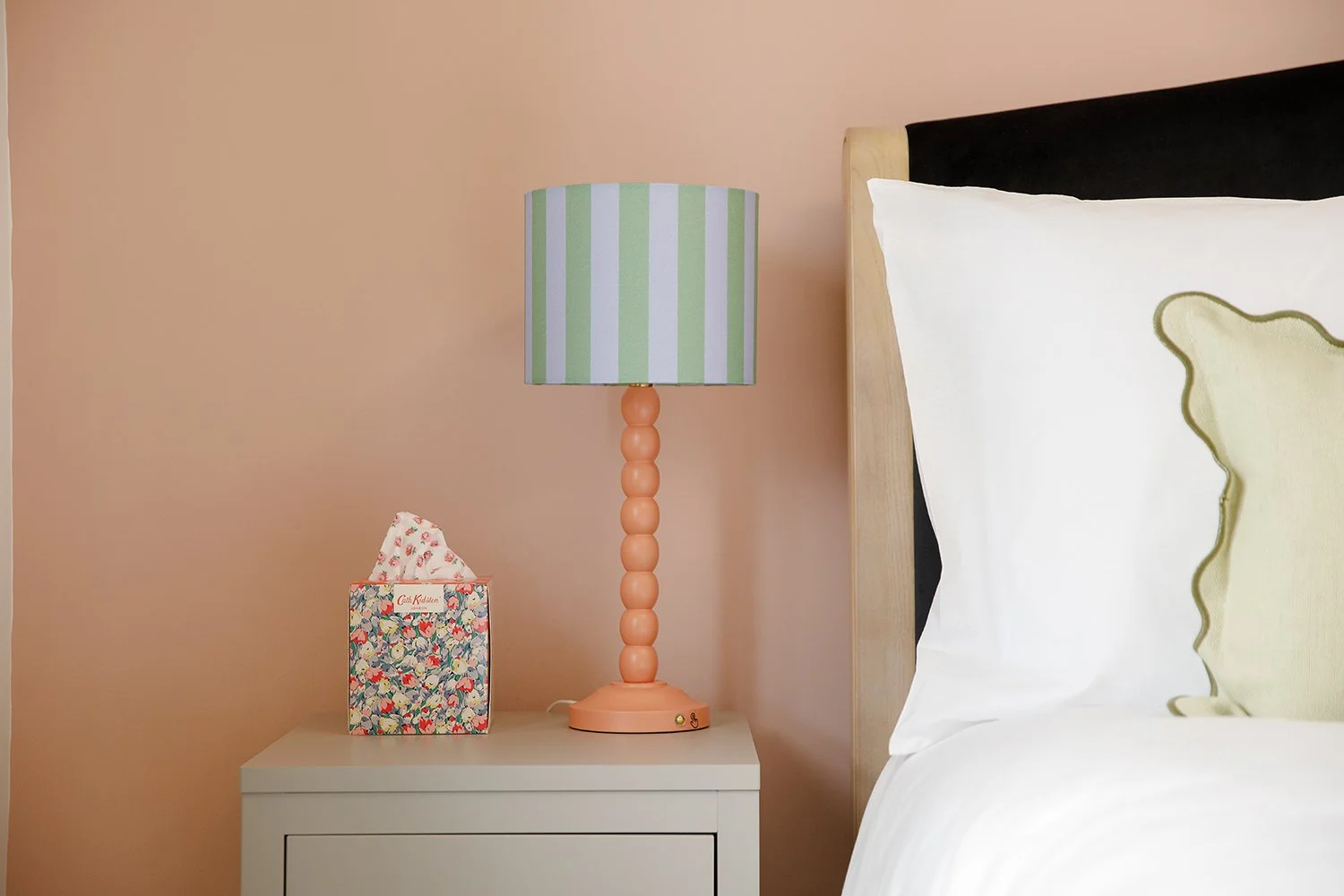 A bedroom nightstand with a colorful floral tissue box and a pink lamp with a striped green and lavender lampshade, next to a bed with white and green pillows against a light peach wall.