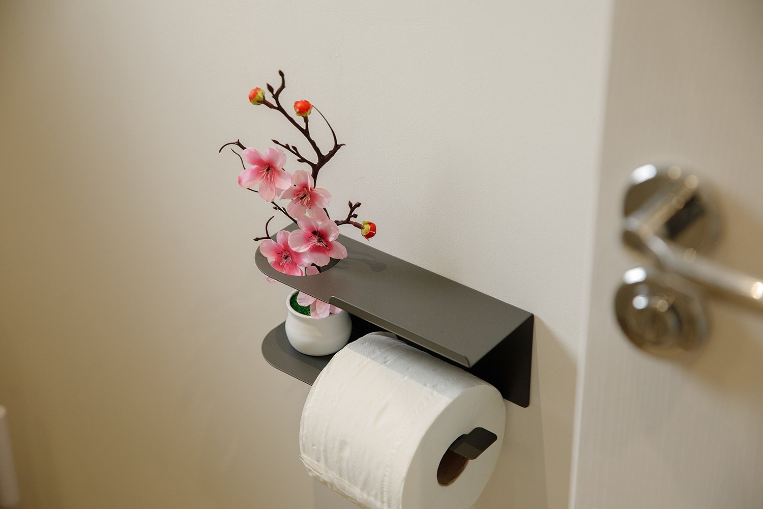A wall-mounted toilet paper holder with a white paper roll, a black shelf holding a pink artificial cherry blossom branch in a white vase, and a white wall behind.