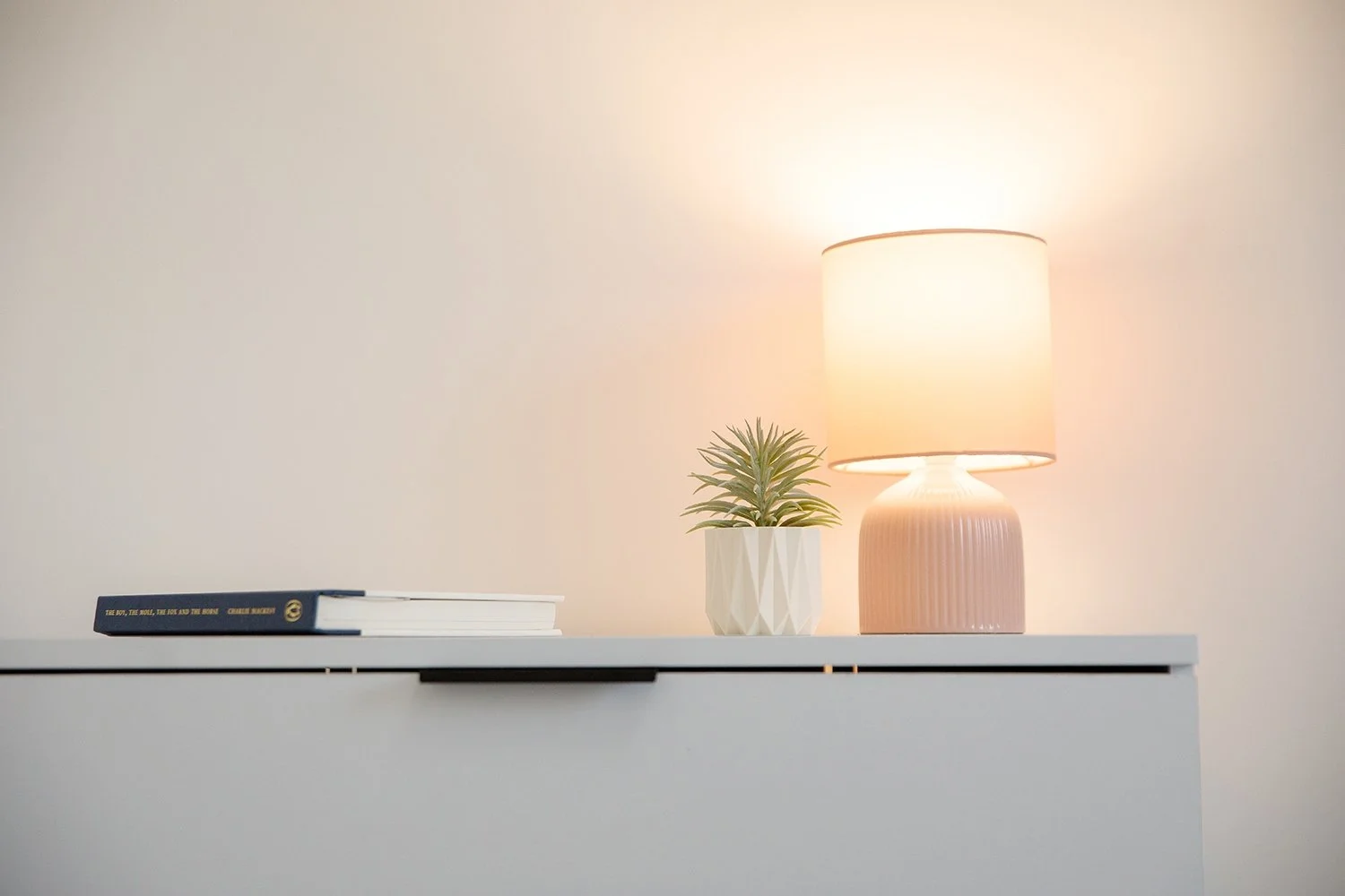 A white tabletop with a small potted plant, a closed book, and a pink lamp emitting warm light against a plain white wall.