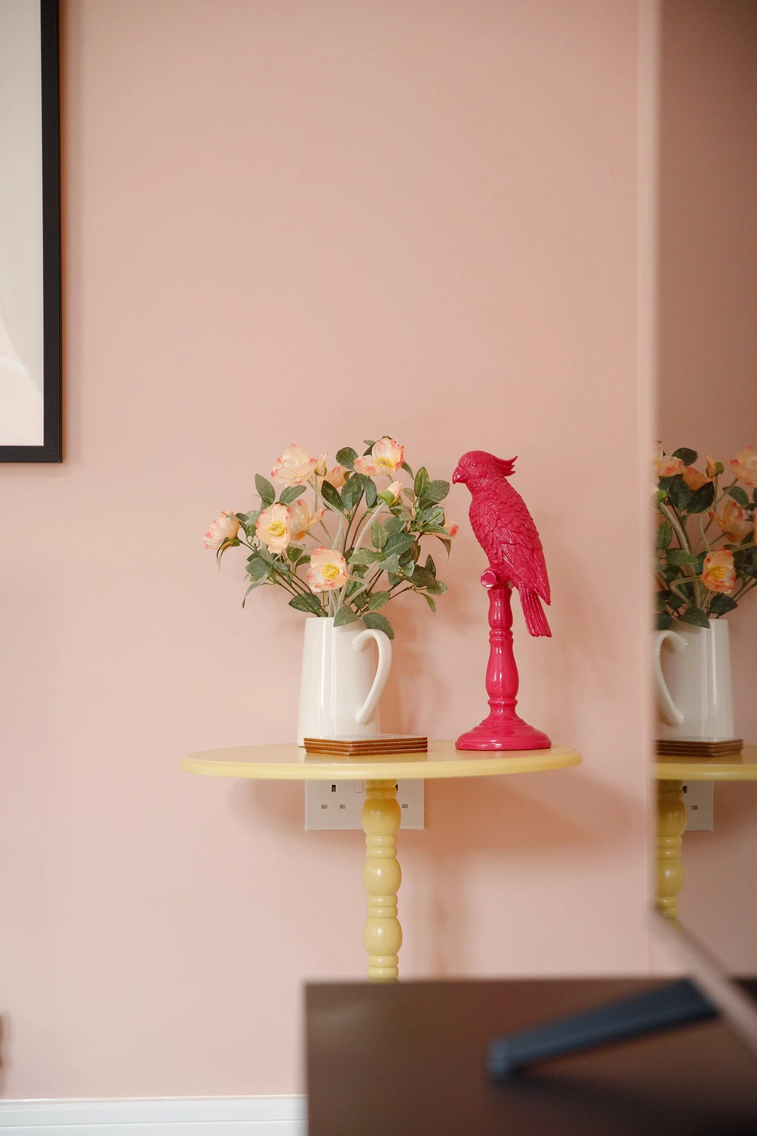 A round yellow table with a yellow turned pedestal base holding a white pitcher with pink and cream artificial flowers and green leaves, and a red bird sculpture on a matching red stand.