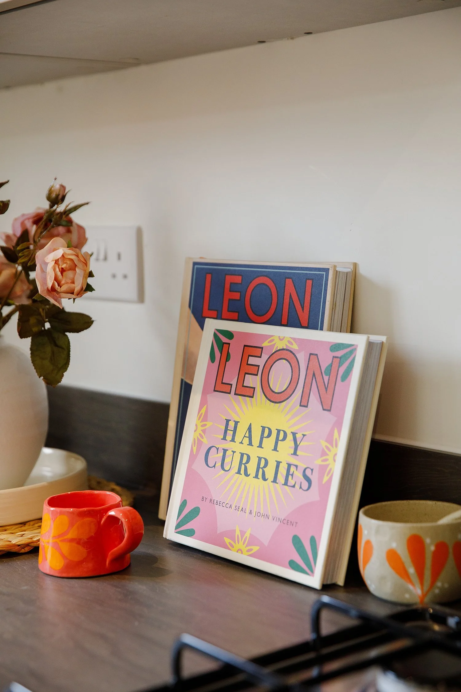 Close-up of a kitchen counter with a pink mug, a decorated bowl, and a pink sign with colorful lettering that reads 'Leon Happy Curie.' There is a vase with pink roses, part of a white wall, and a cookbook or magazine with a pink cover next to the si