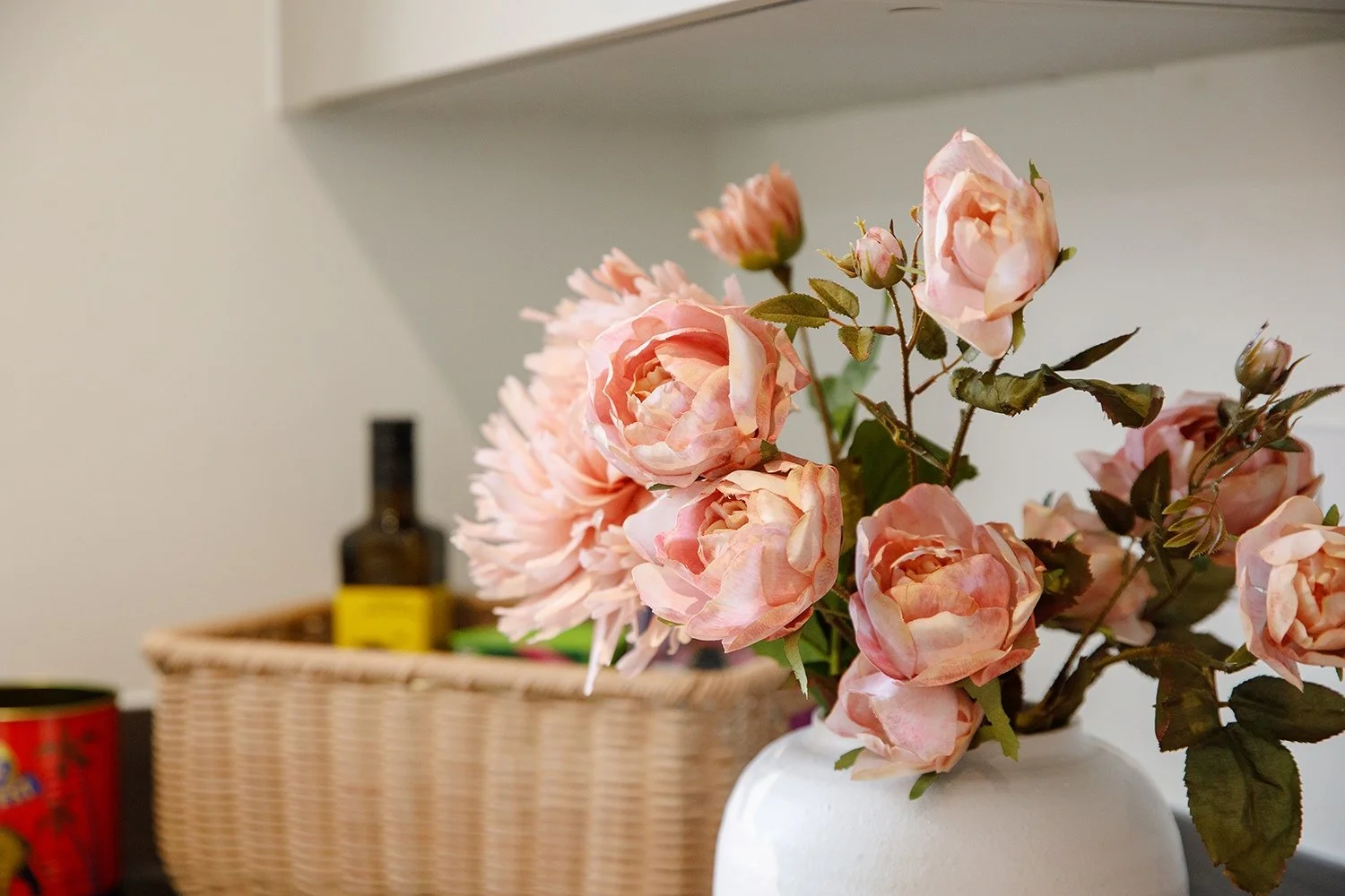 Pink artificial roses in a white vase with a basket and bottles in the background.