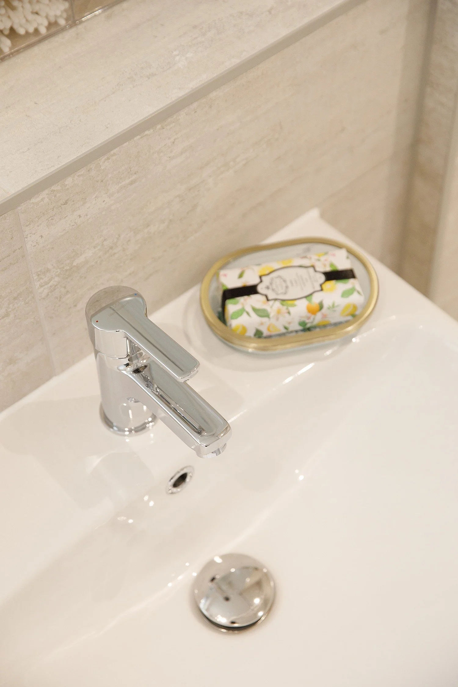 A bathroom sink with a chrome faucet and a soap holder. A rectangular soap or packet with a floral pattern and a black band is inside the soap holder. The background shows beige tiled wall and a countertop.