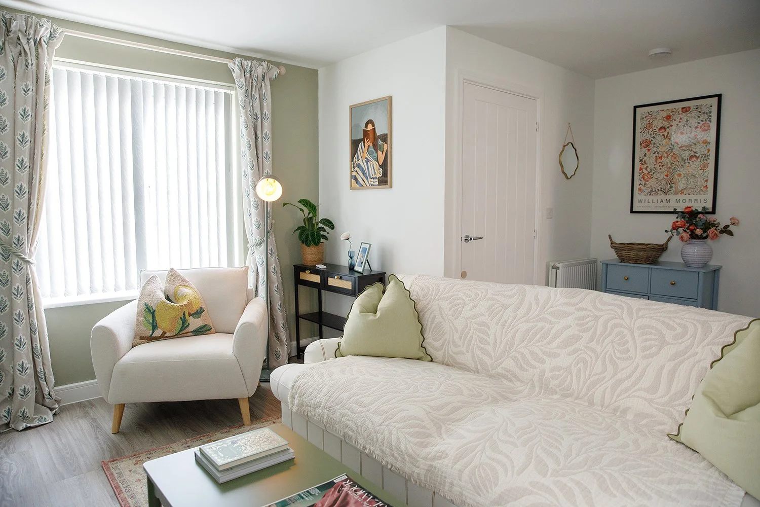A cozy living room with a white armchair and a sofa, a window with patterned curtains, a small black side table with a potted plant and framed photos, a blue cabinet with a flower vase, and various wall art, including a William Morris print