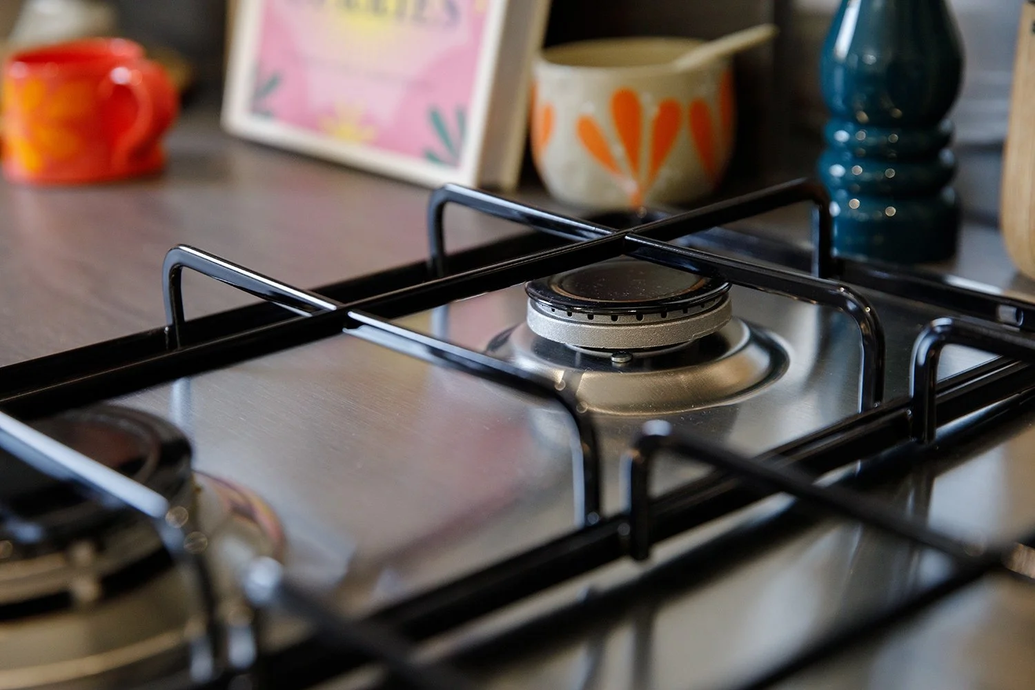 Close-up of a gas stove burner on a kitchen countertop with decorative cups, framed artwork, and vases in the background.