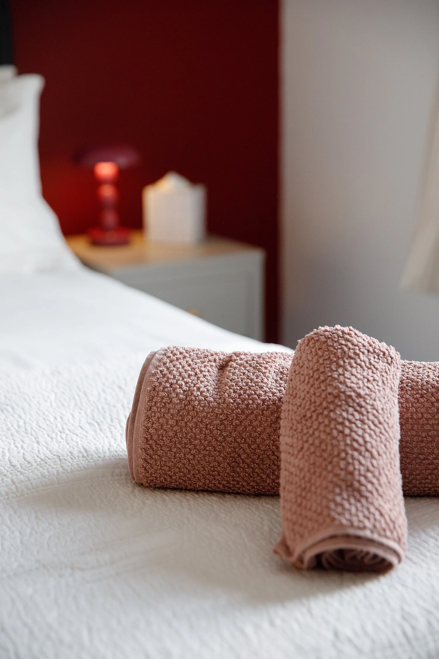 Rolled pink towel on a white bed with a blurred red lamp and white candle in the background.