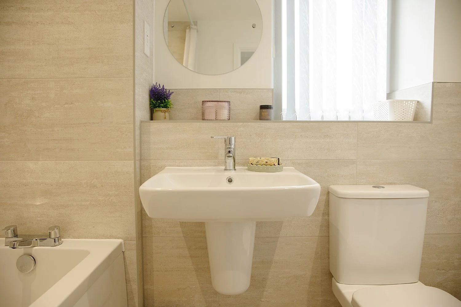 A modern bathroom with a white sink, toilet, and partial bathtub, beige tiled walls, round mirror, and a window with vertical blinds. Decor includes a potted plant, candle, soap, and small basket.