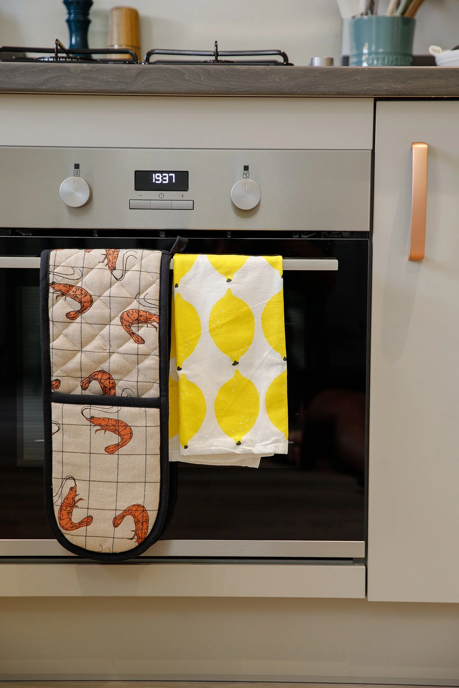 Kitchen oven with two oven mitts hanging on the handle, one with shrimp design and the other with lemon pattern, and a digital clock displaying 19:37.