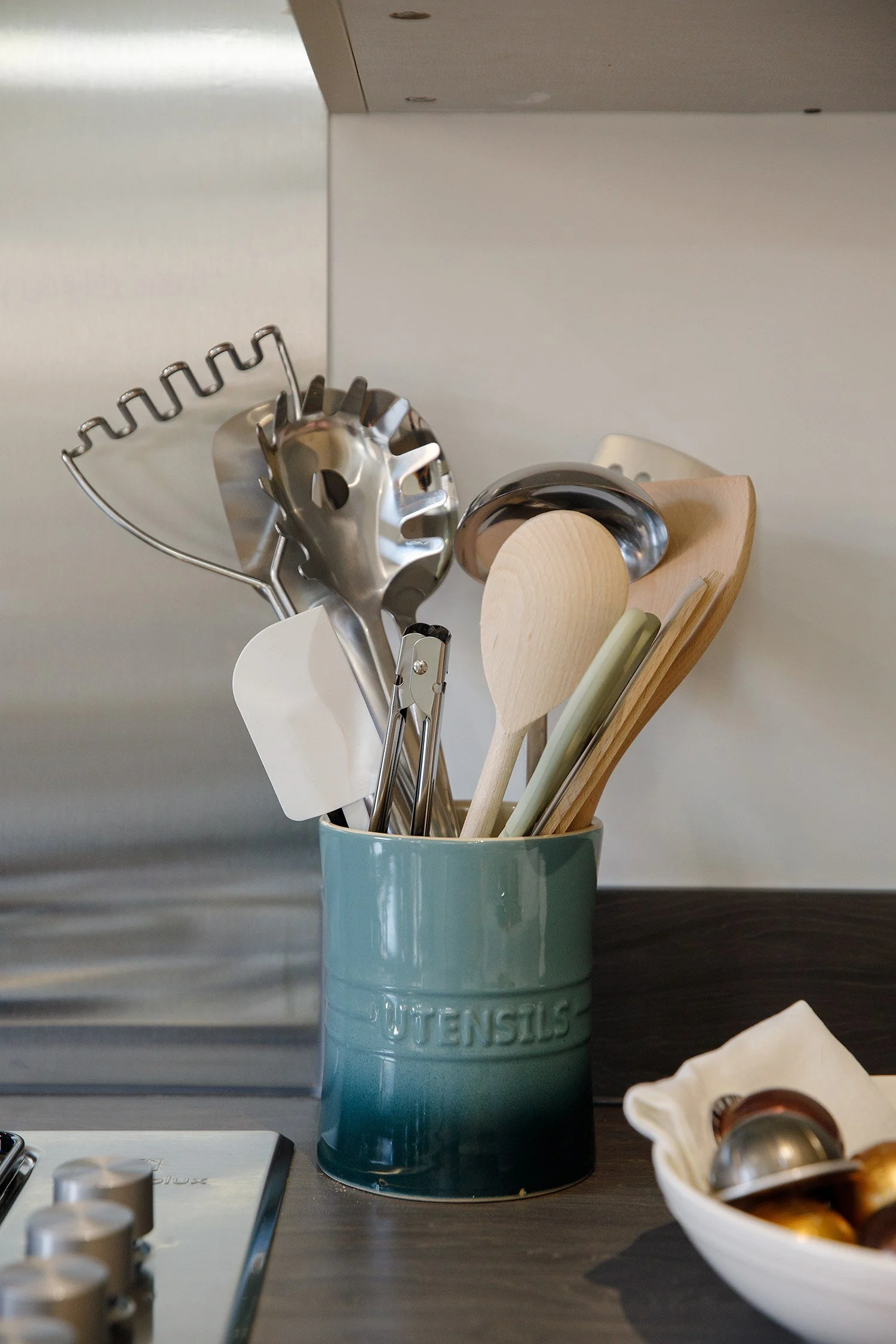 A blue kitchen utensil holder with the word "UTENSILS" embossed on it holds various kitchen tools like a metal whisk and a wooden spoon. The holder is on a dark kitchen counter with a white bowl containing metallic objects and a partial view of a gas