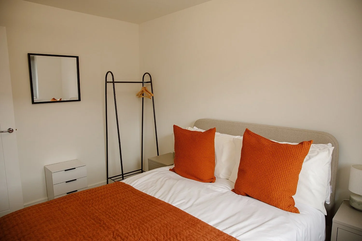 A cozy bedroom with a bed featuring white sheets, orange pillows, and an orange quilt. A nightstand with a small lamp is beside the bed, and a wall-mounted mirror and a black metal clothes rack are also visible.