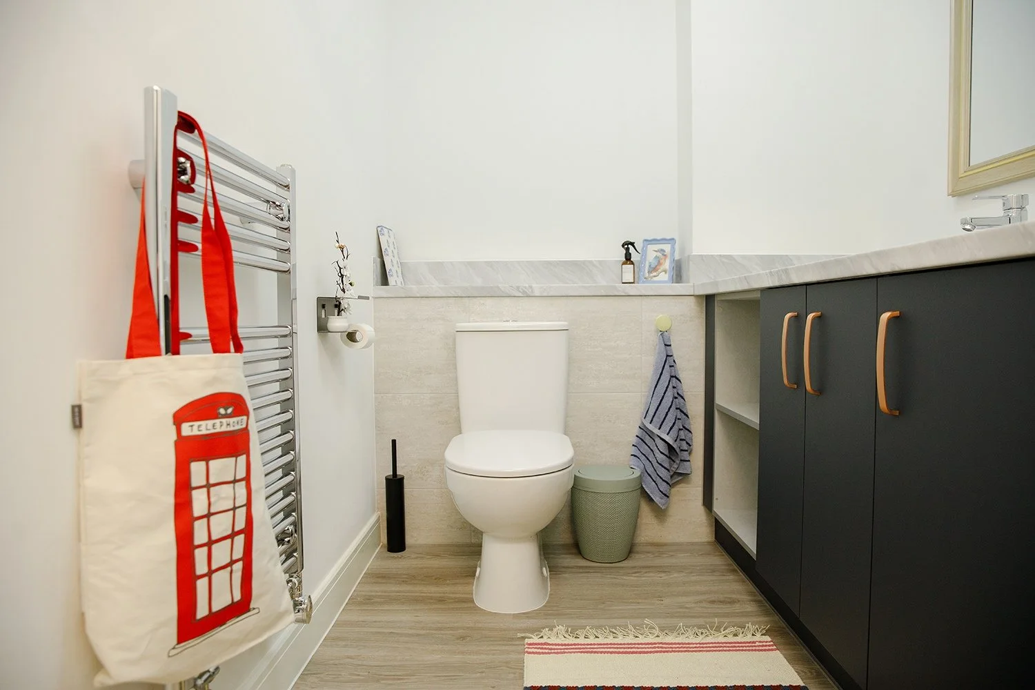 A small bathroom with a toilet, a dark gray cabinet, a gray waste bin, and a towel hanging on the wall. A towel rack with a tote bag that has a red telephone booth design is mounted on the wall. The bathroom has a marble countertop with a mirror abov
