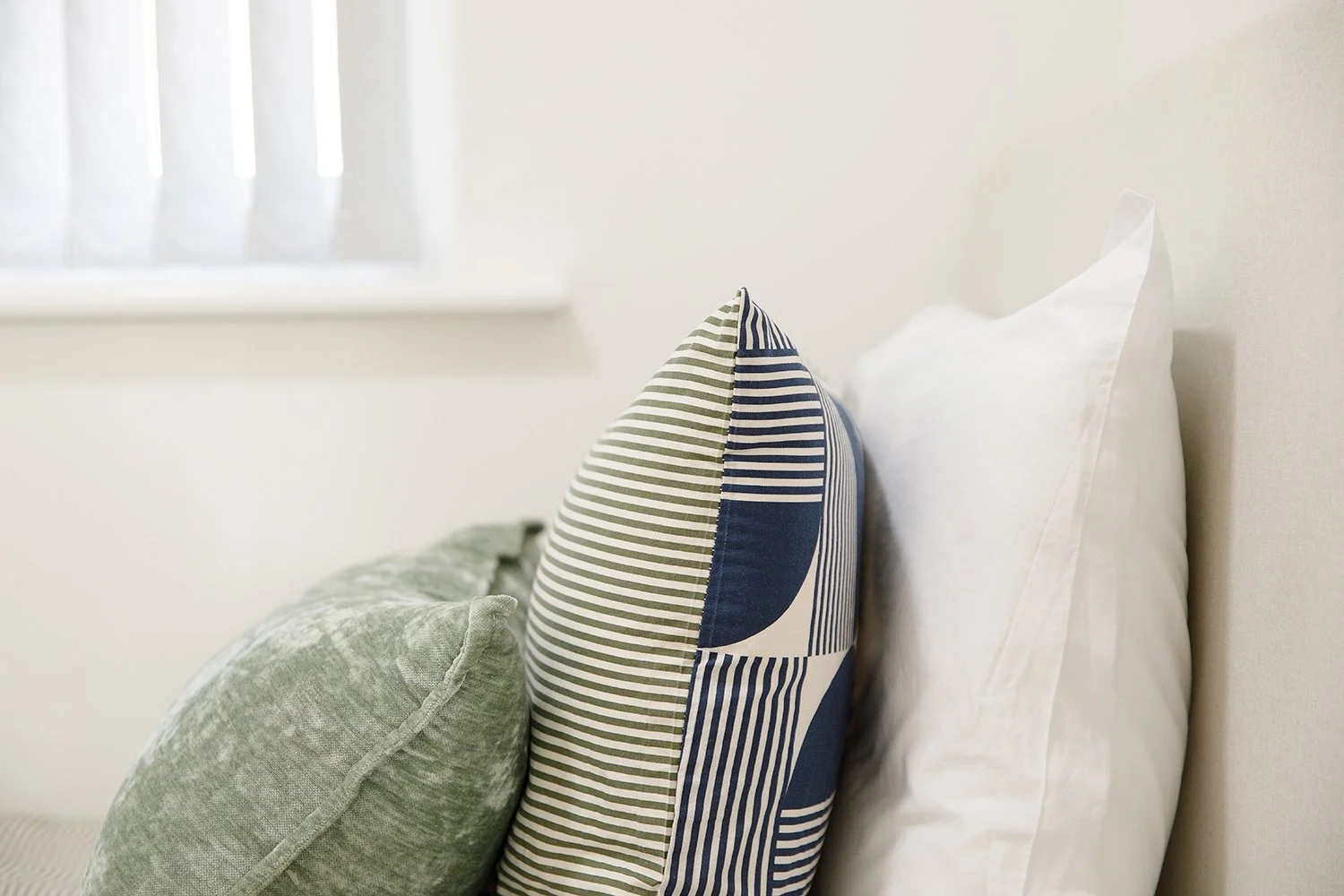 Close-up of a bed with multiple pillows, including a white pillow, a textured green pillow, and a blue and white striped pillow, in a well-lit room.