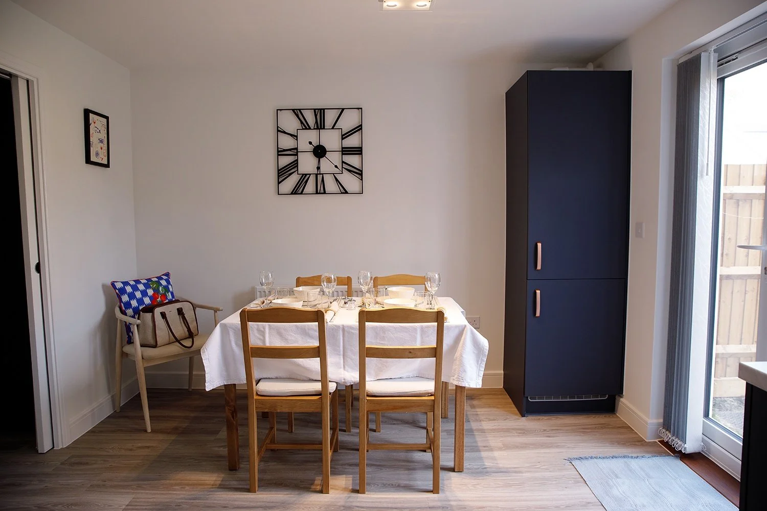 Dining room with a rectangular table set with wine glasses and napkins, four wooden chairs, a black cabinet, a wall clock, a small framed artwork, and a sliding glass door with vertical blinds leading to an outdoor area.