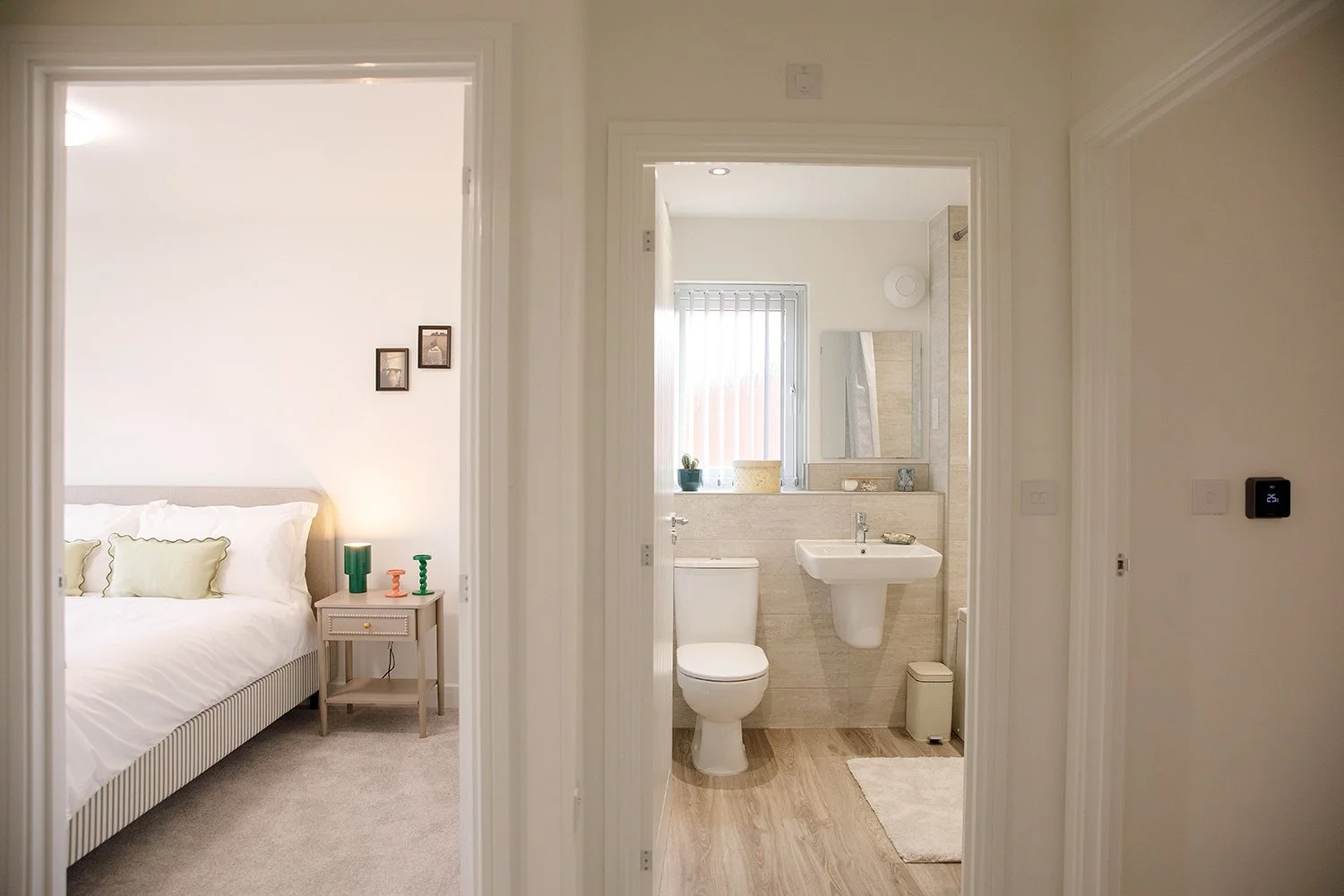 View of a bedroom on the left with a bed, nightstand, and decorative pillows, and a bathroom on the right with a toilet, sink, mirror, and window with vertical blinds.