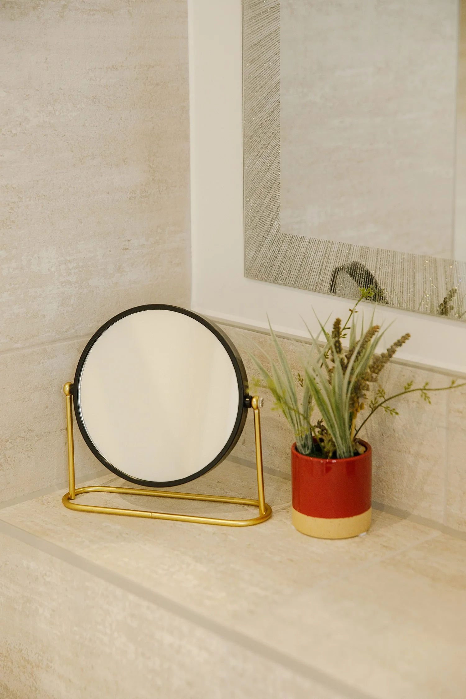Decorative mirror and potted plant on beige countertop, with beige wall and framed artwork in the background.