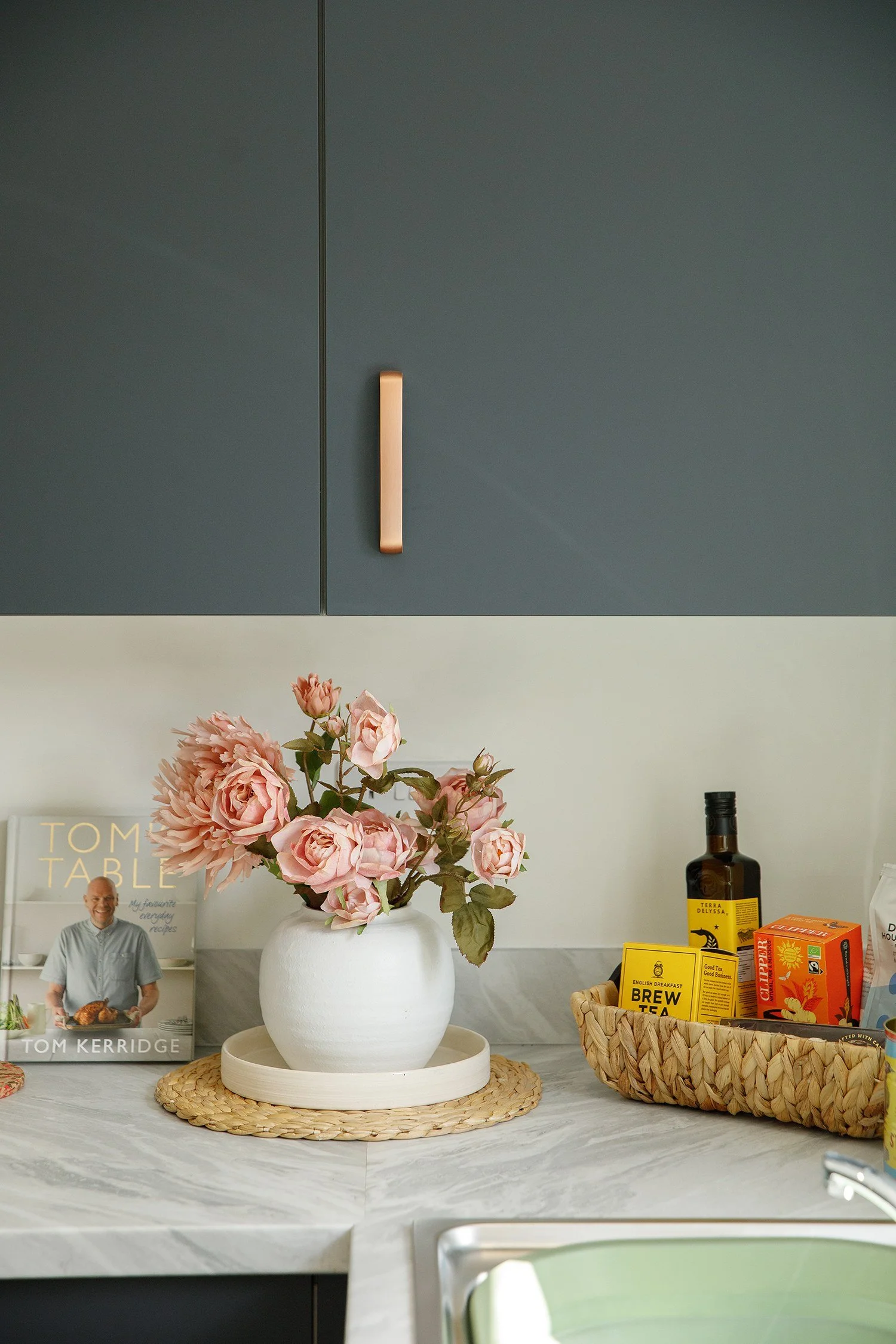 A white vase with pink flowers on a round woven placemat on a marble kitchen countertop. In the background, there are kitchen cabinets, a selection of tea and food items, a magazine featuring Tom Kerridge, and a man with a beard and glasses holding a