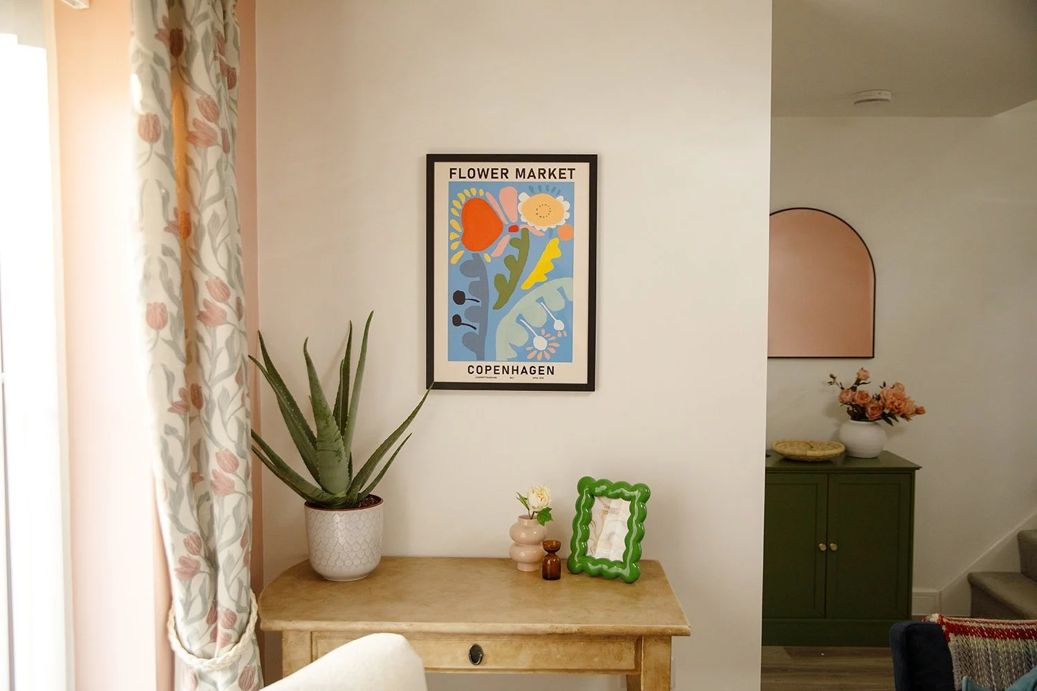 Decorative interior space with a framed poster of a flower market in Copenhagen on the wall, a potted aloe vera on a wooden console table, small vases with flowers, and a green decorative mirror. Part of a window with patterned curtains and a glimpse