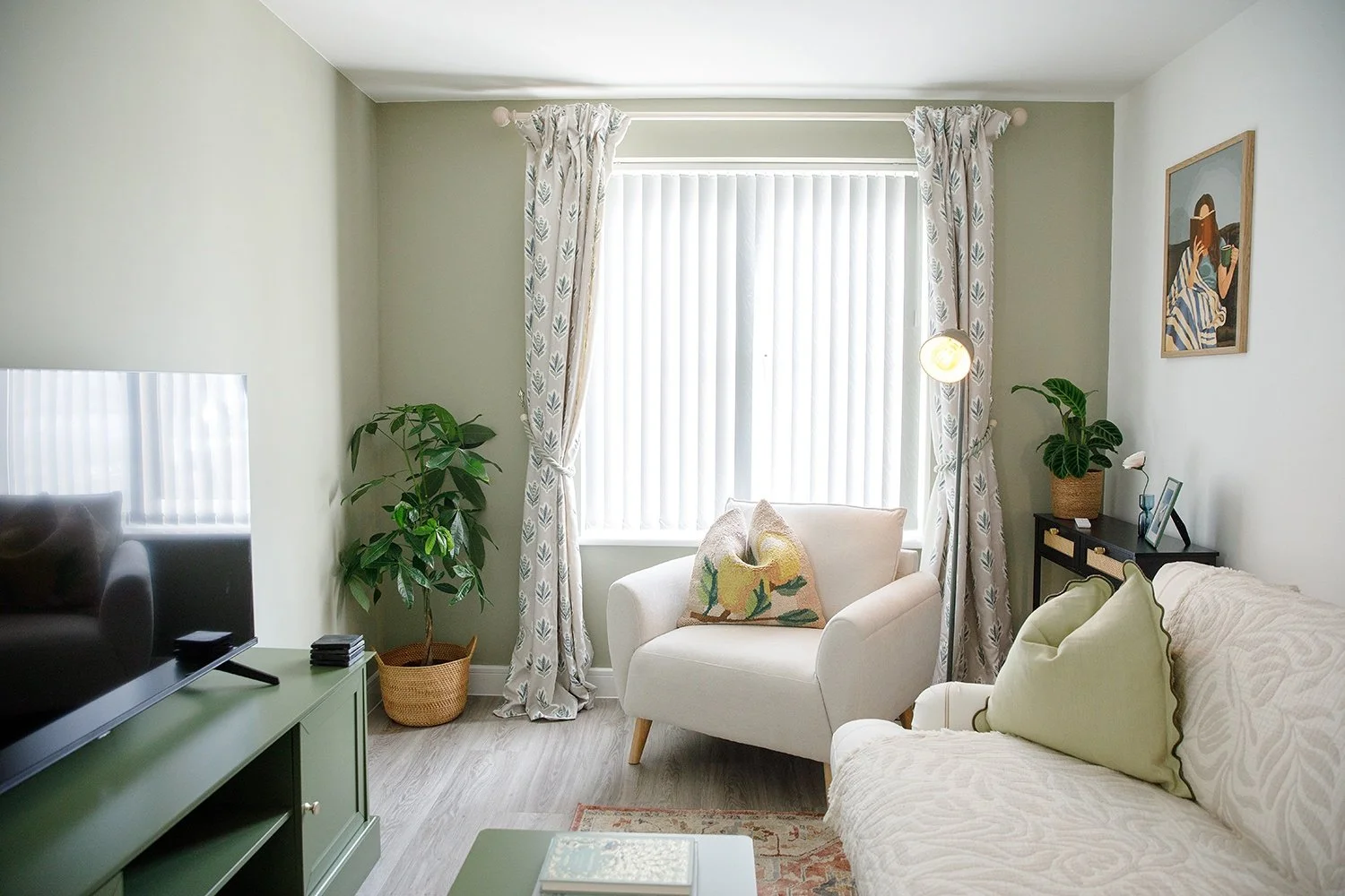 Living room with a white armchair, a green TV stand, a flat-screen TV, potted plants, a sofa with a pillow, curtains, a wall picture, and a side table.