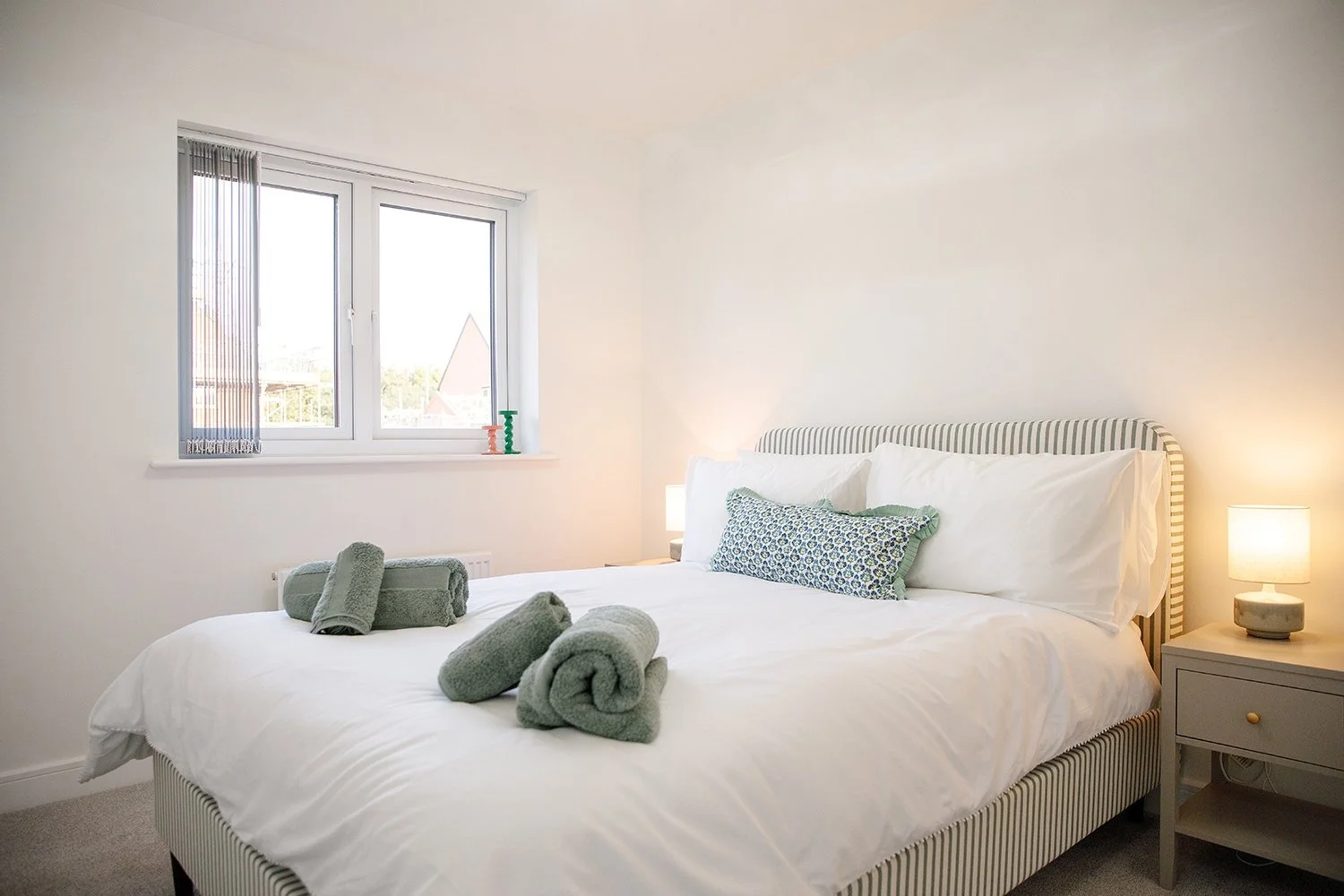 A neatly made bed with white sheets and multiple pillows, topped with rolled gray towels, in a bright bedroom with a window, bedside lamp, and a gray nightstand.