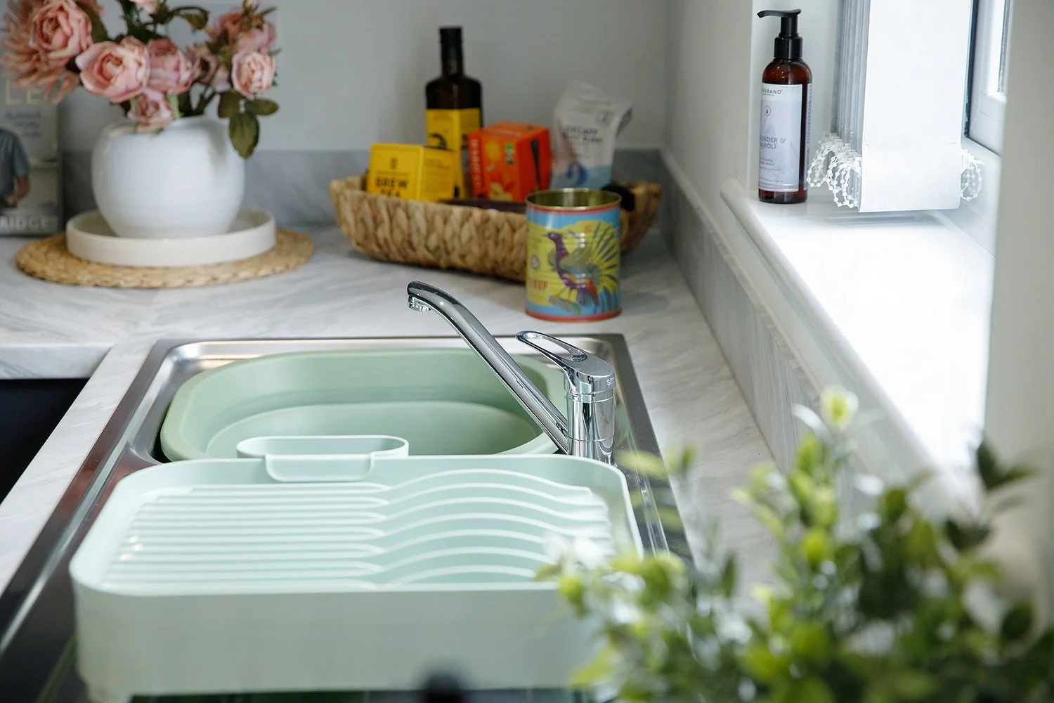 Kitchen sink with a chrome faucet, a green dish drying rack, placed on a marble countertop near a window. In the background, a basket with various boxes, a glass bottle, a colorful mug, and a white vase with pink flowers are visible.