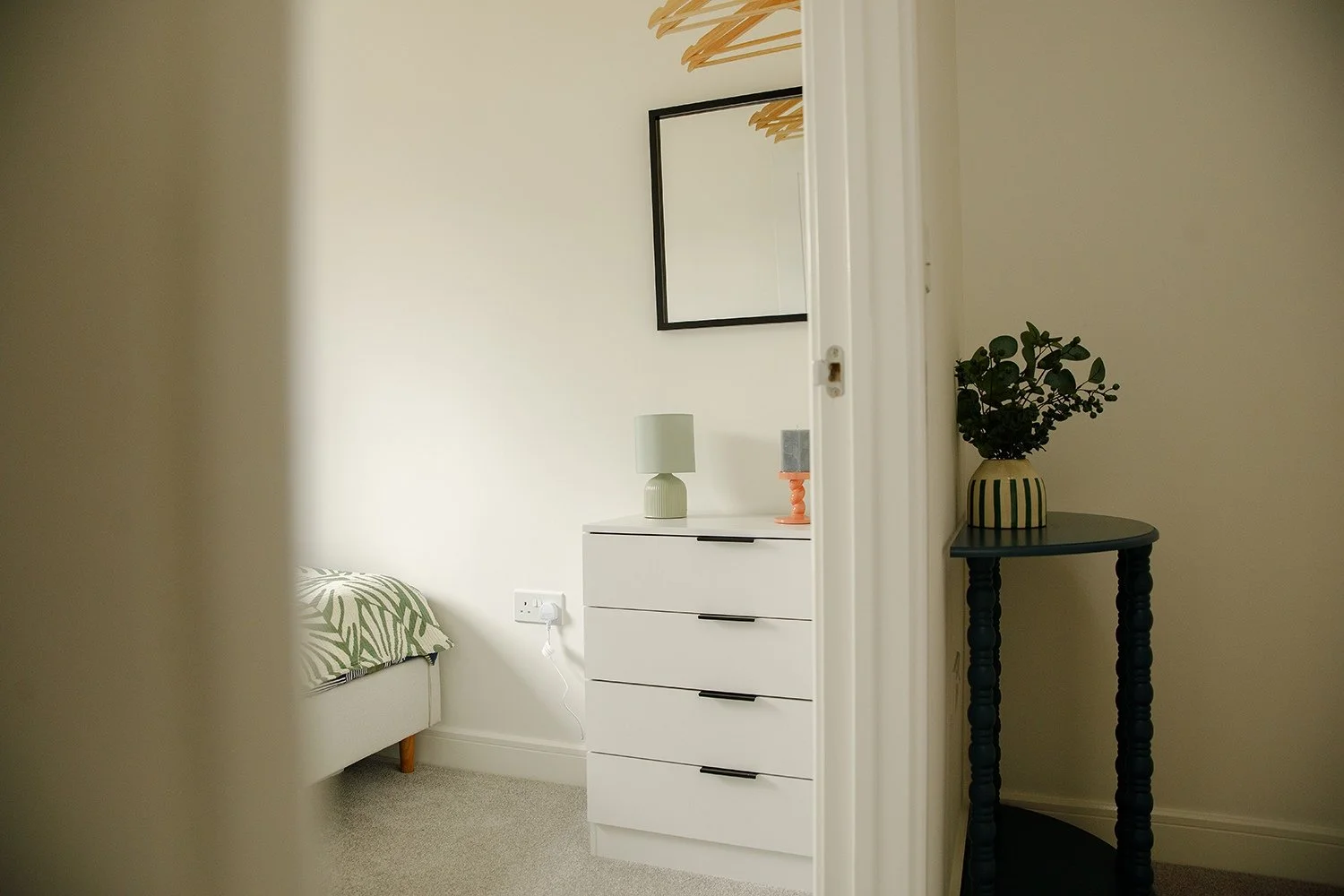 Partially visible bedroom with a bed and green patterned pillow, white dresser with a small lamp, mirror on the wall, and a black side table with a potted plant.