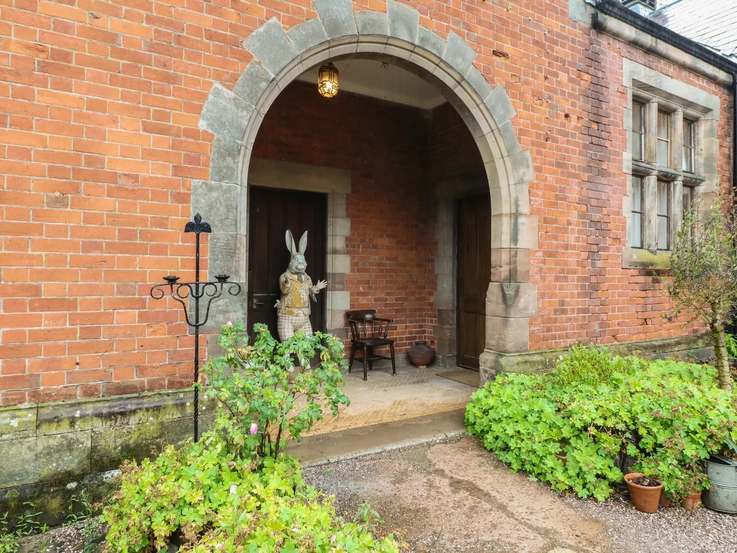 The entrance of a brick house with a rounded stone archway. There is a decorative rabbit statue dressed in clothing, waving. There are potted plants, a wooden chair, and an iron candle holder near the doorway.