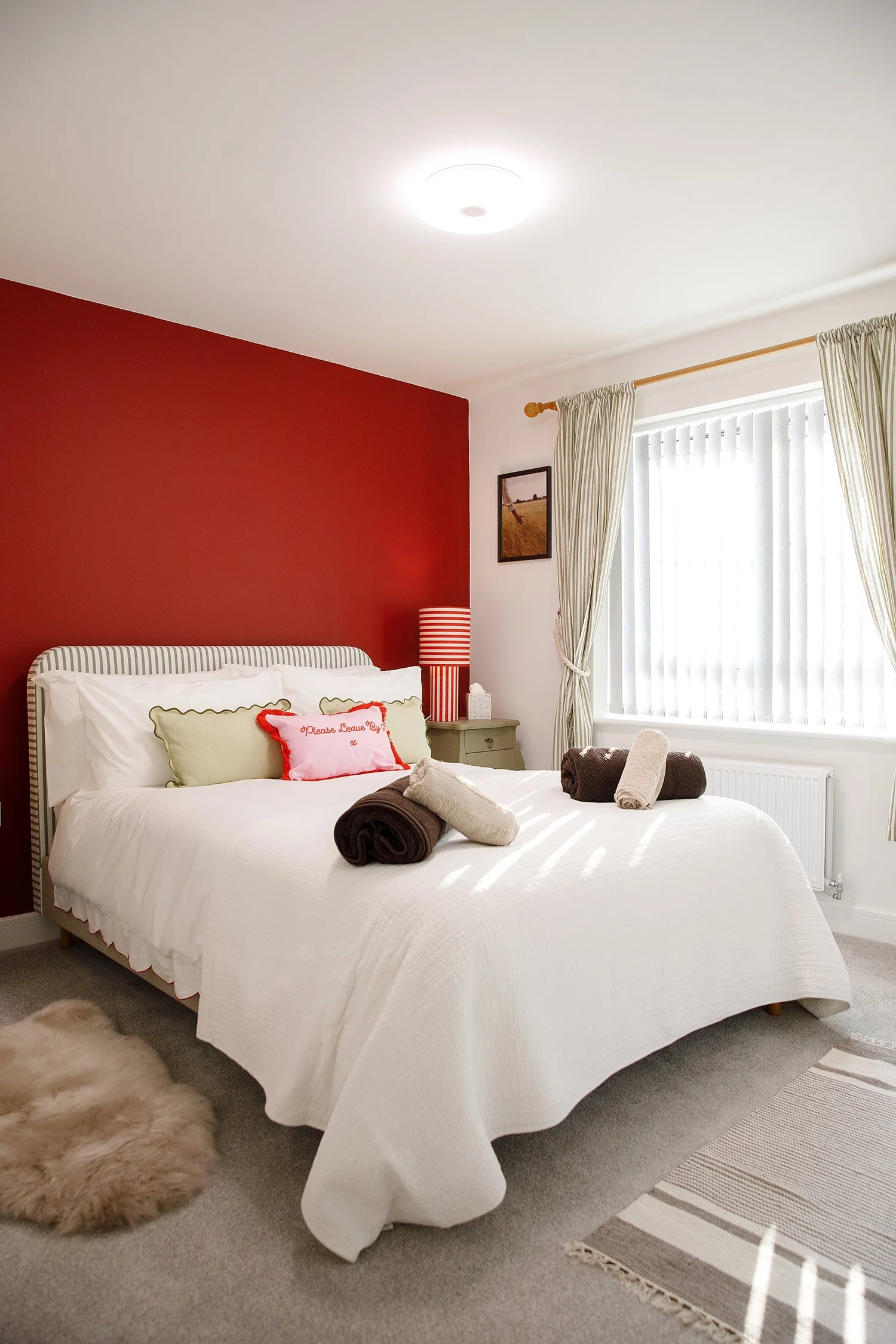 A neatly made bed with a white quilt, decorative pillows, and rolled towels on top. There is a red accent wall behind the bed, a small gray nightstand with a striped red and white lamp, a framed picture on the wall, and a window with cream-colored cu
