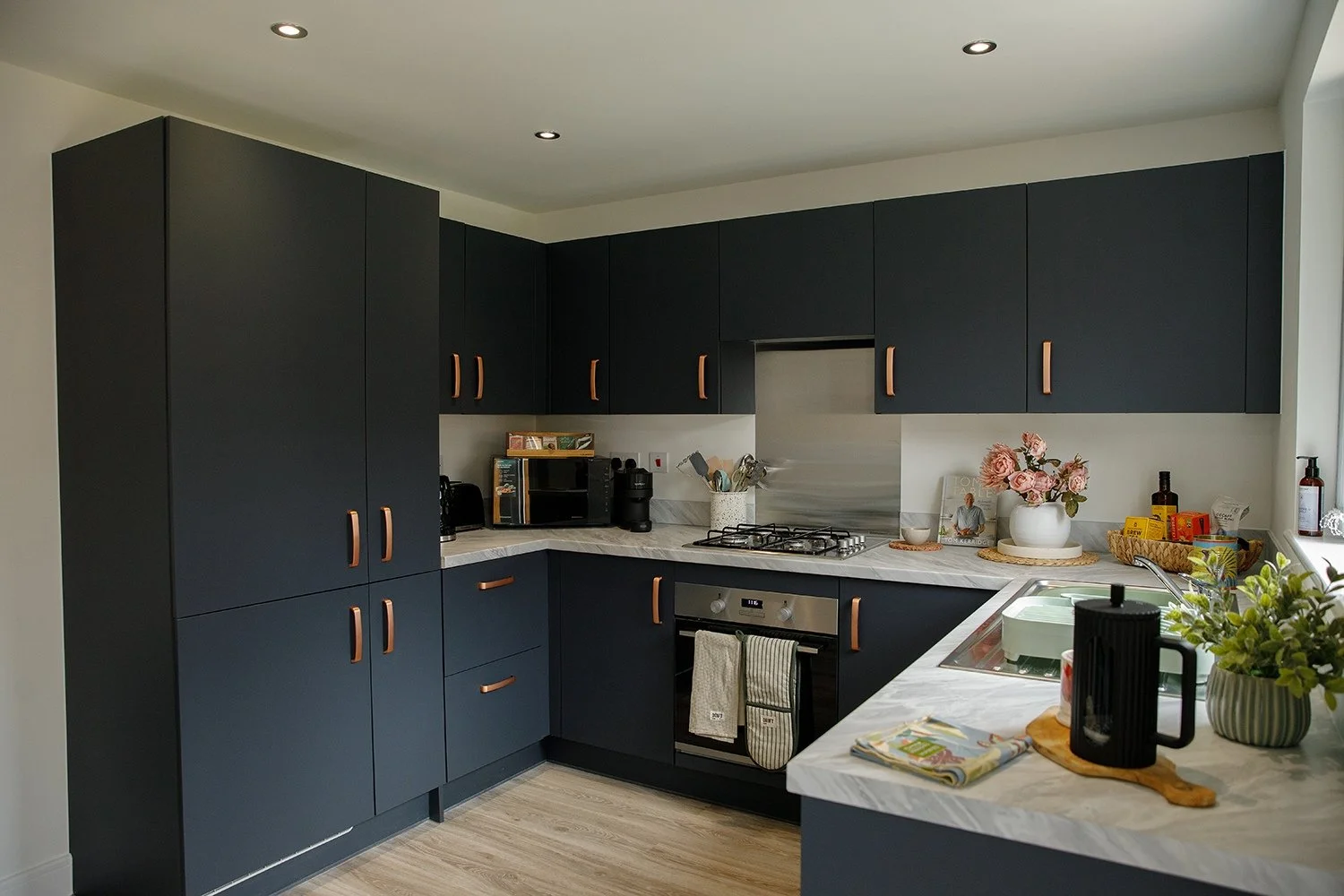 Modern kitchen with black cabinets, marble countertops, stainless steel appliances, and decor including a bouquet of pink roses, potted plants, and kitchen utensils.