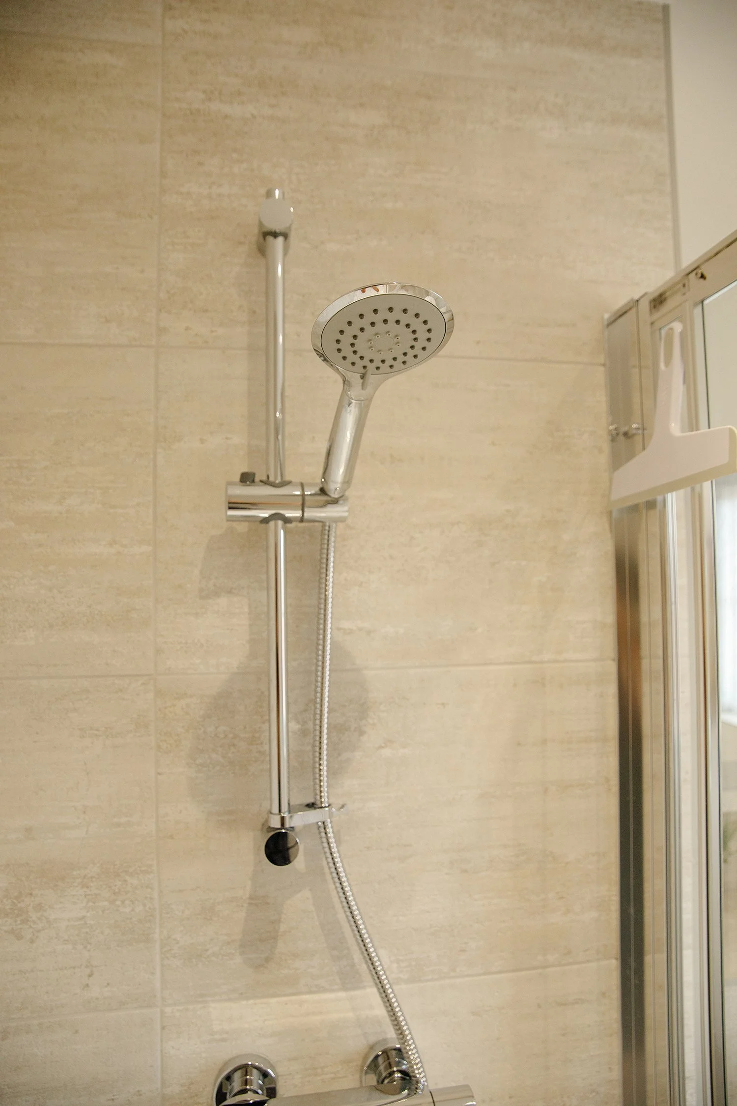 A modern showerhead attached to a beige tiled wall with a handheld shower hose, and a shower control panel below.