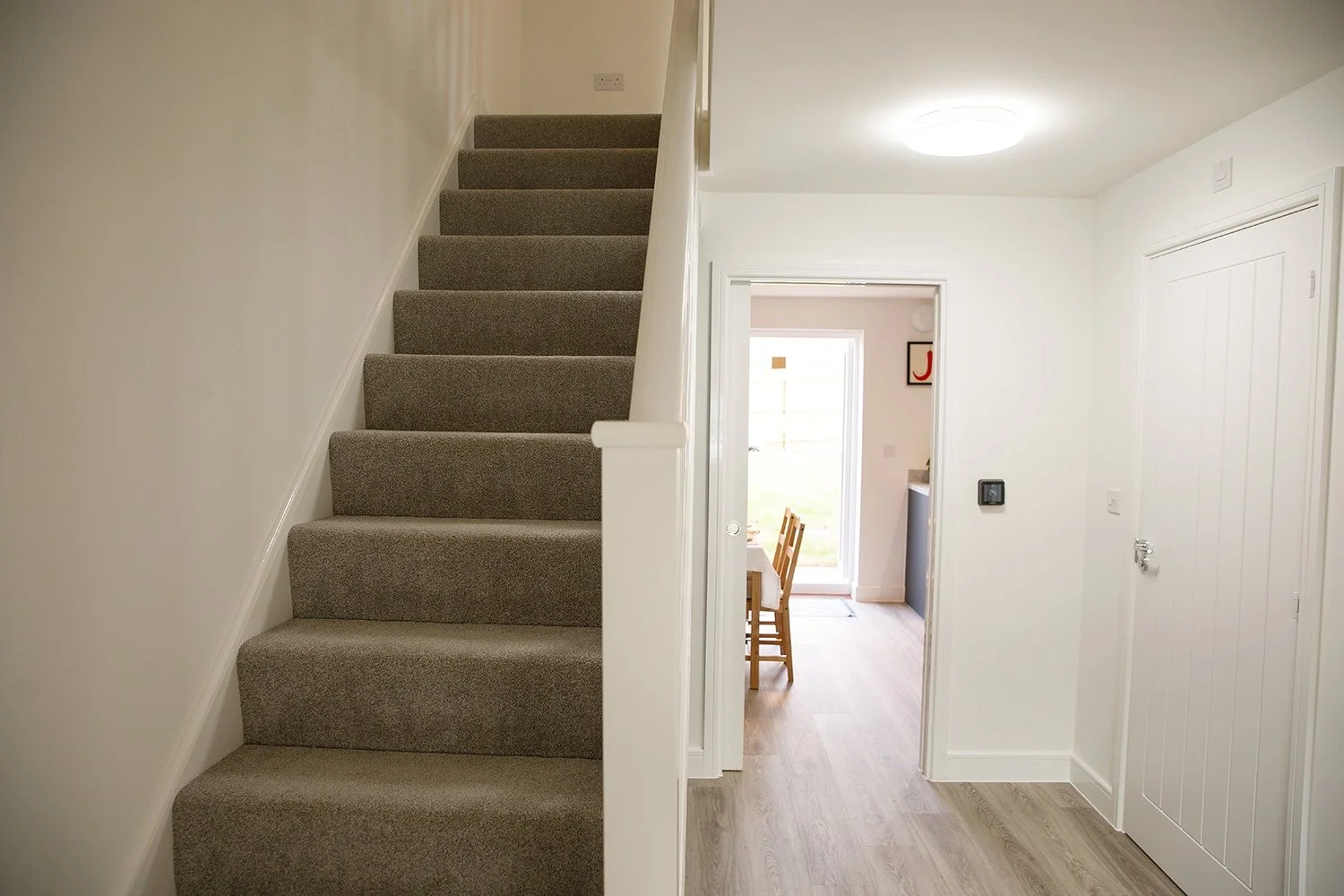 Interior view of a home with beige carpeted stairs on the left, white walls, and a doorway leading to a dining area with chairs and a glass door in the background.