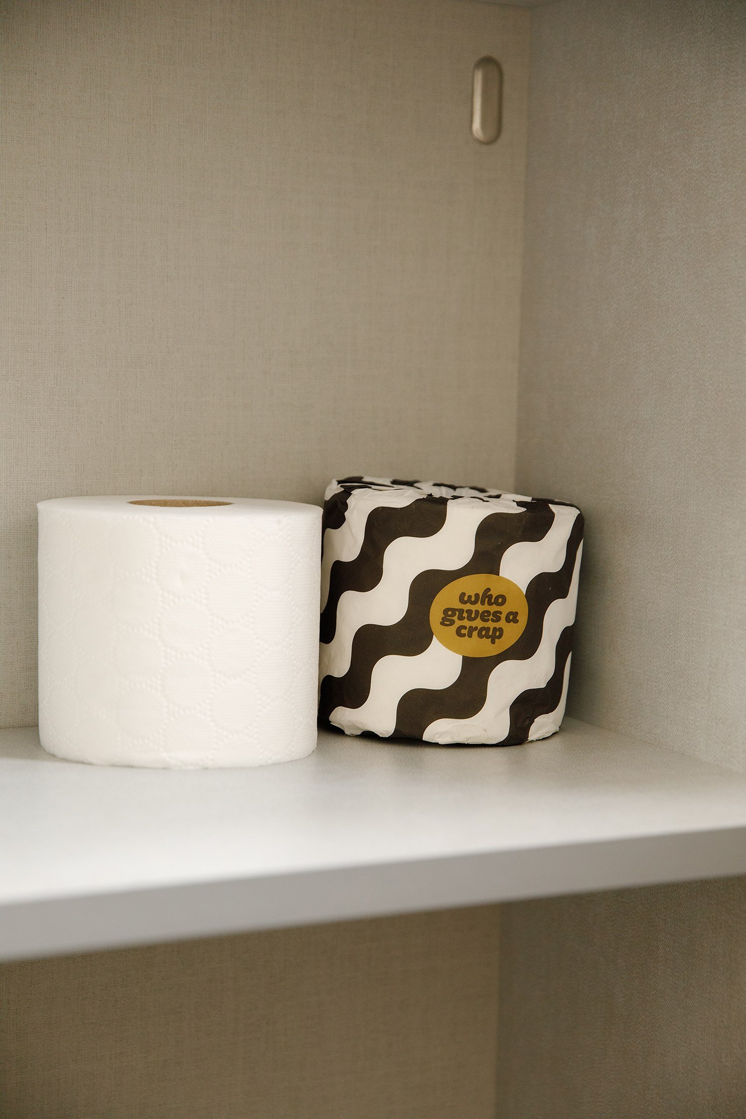 A roll of white toilet paper and a gift bag with black and white wavy pattern and a yellow circle with the words 'who gives a crap' on a white shelf against a beige wall.