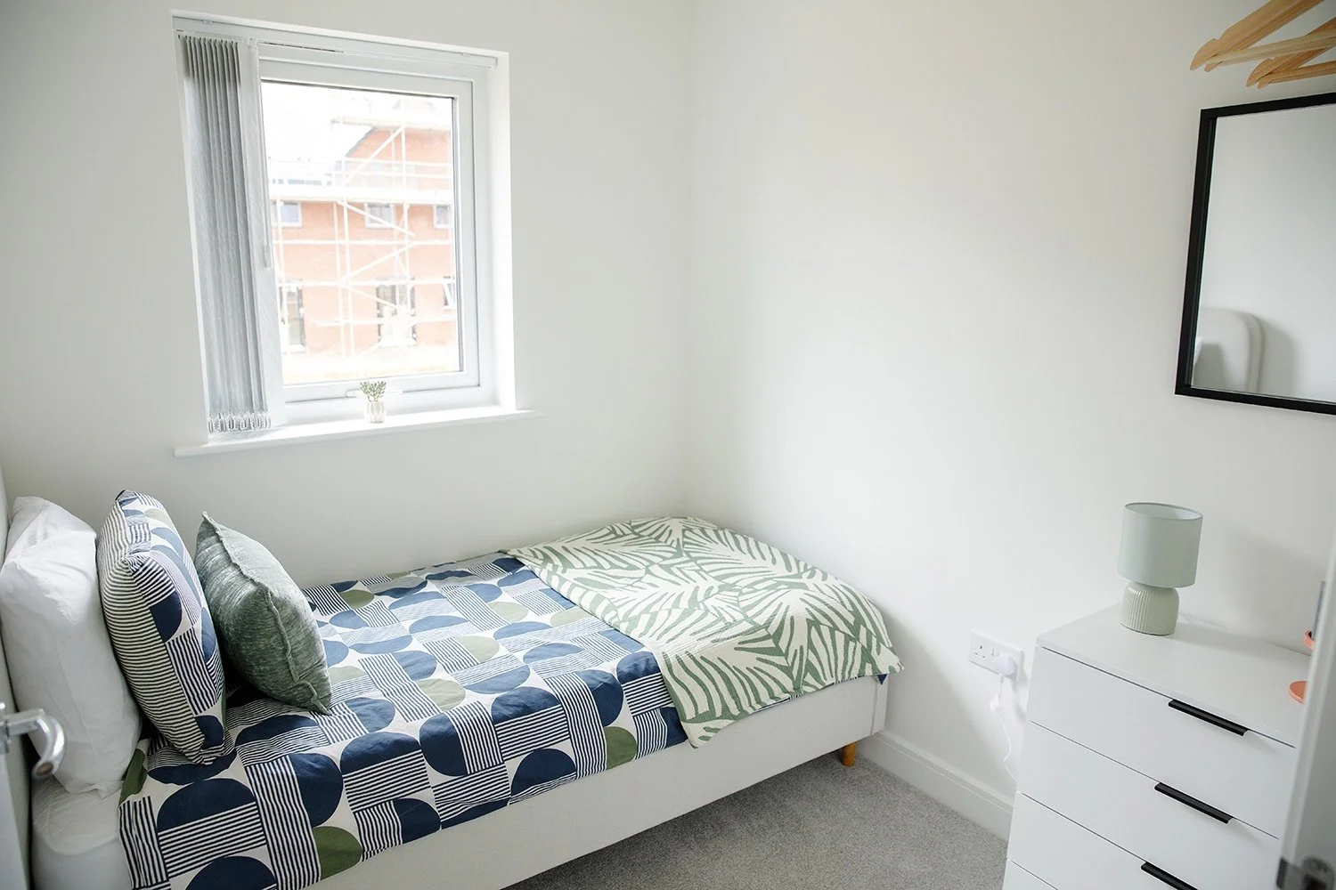 A small bedroom with a single bed, decorated with patterned pillows and bedspread, a window with vertical blinds, and a white dresser with a lamp and mirror.