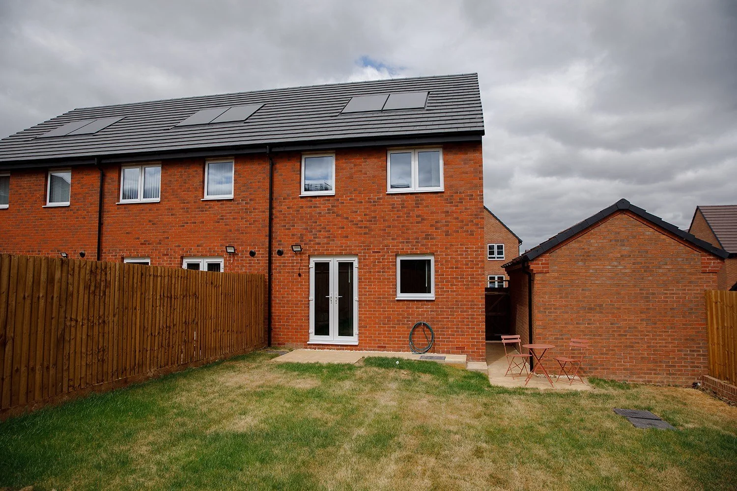 Backyard view of a modern brick house with solar panels on the roof, a wooden fence, a small patio with pink outdoor furniture, and a grassy lawn.
