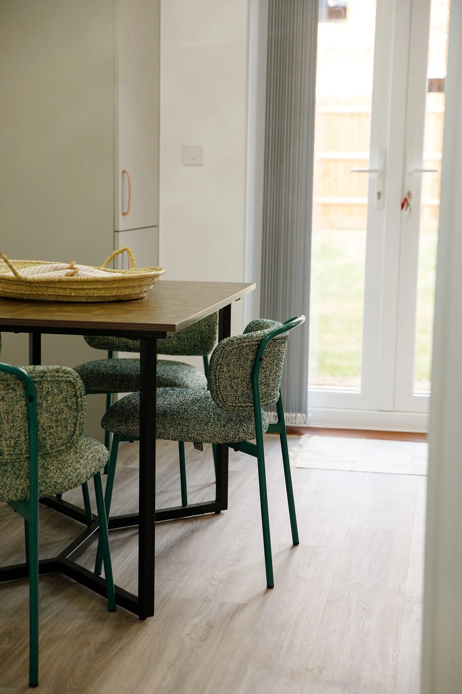 A wooden dining table surrounded by four green patterned chairs in a room with light-colored wood flooring and a sliding glass door leading outside.