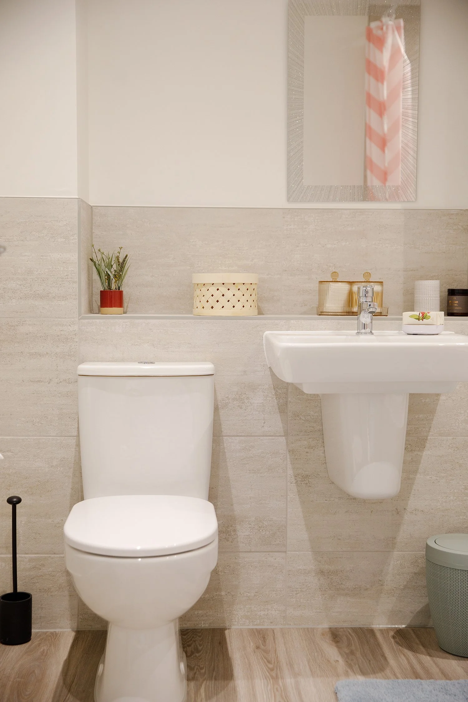 A clean, modern bathroom with a white toilet, a wall-mounted sink with a chrome faucet, a mirror, and decorative items such as a small potted plant, a woven storage basket, and candles. The room features wood-like flooring and beige tiled walls.