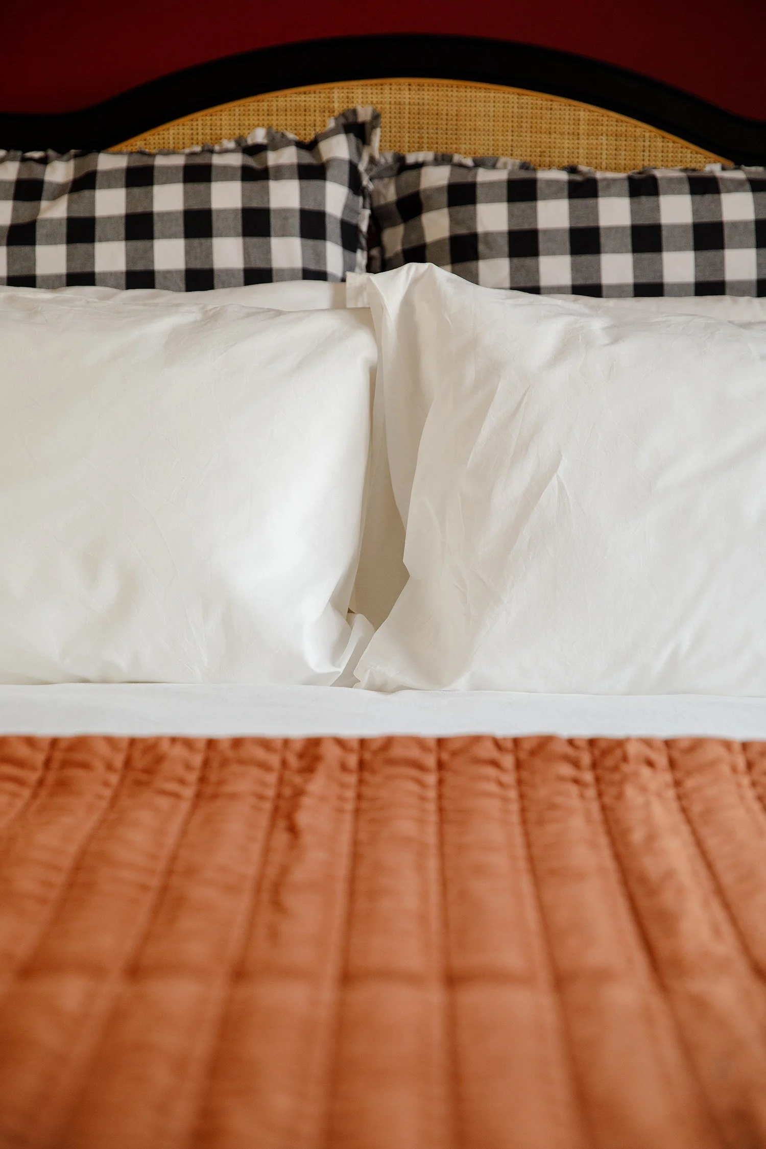 A bed with white sheets, black and white checkered pillows, a rattan headboard, and a reddish-brown wall in the background.