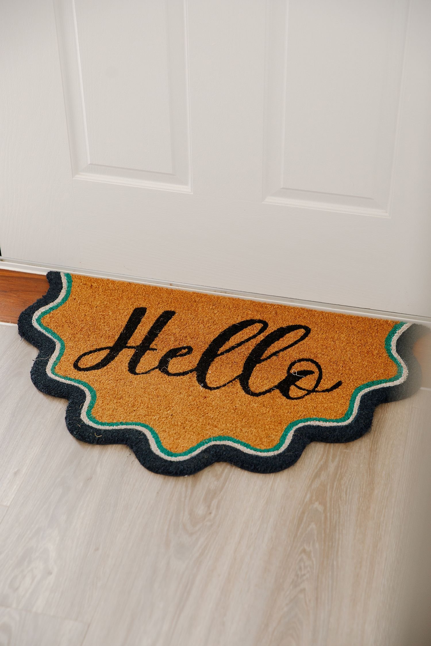 A woven doormat with the word 'Hello' written in black cursive letters on an orange background, trimmed with a teal and black border, placed on a wooden floor in front of a white door.