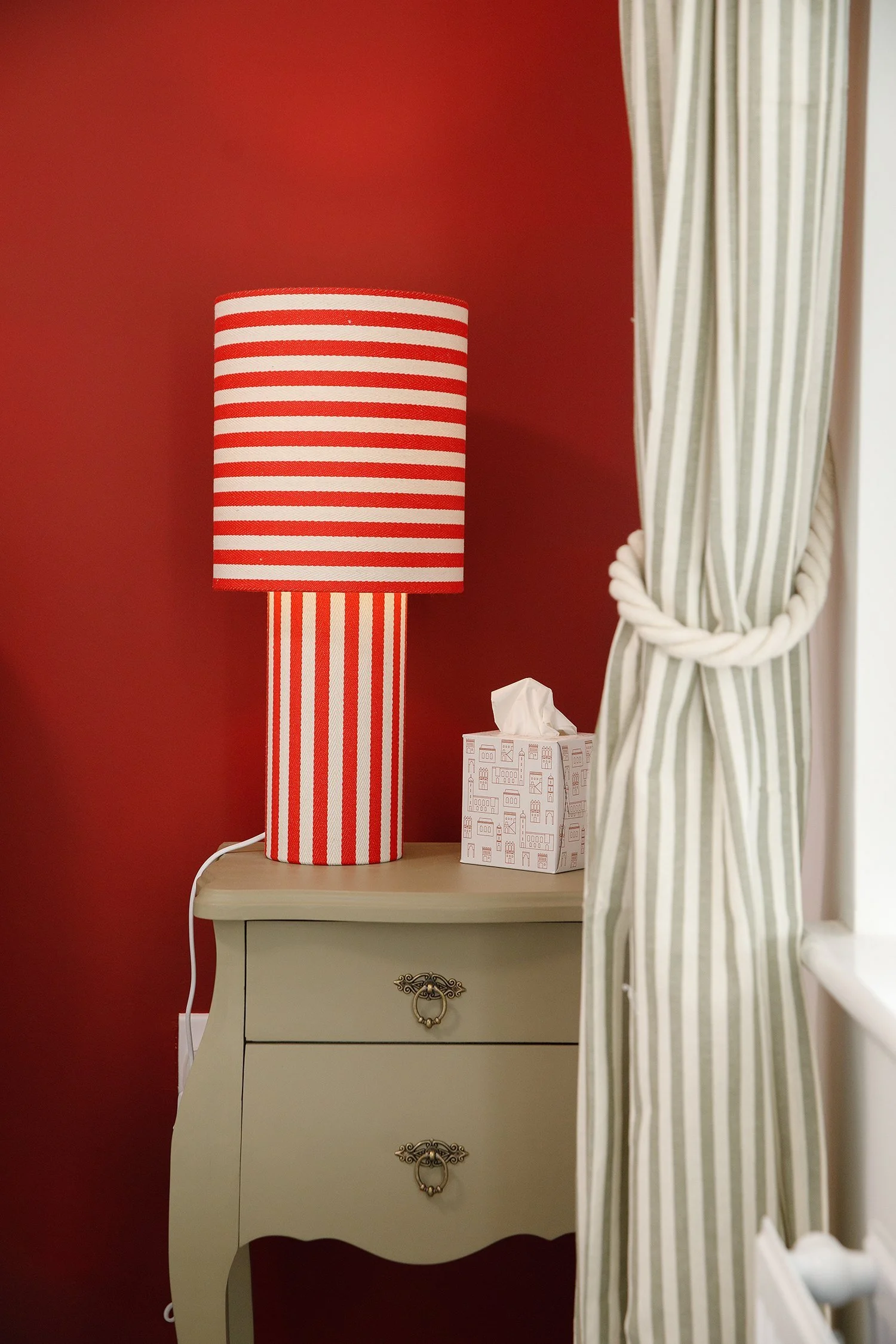 A red and white striped table lamp on a cream-colored nightstand, with a tissue box and a window with striped curtains in the background.