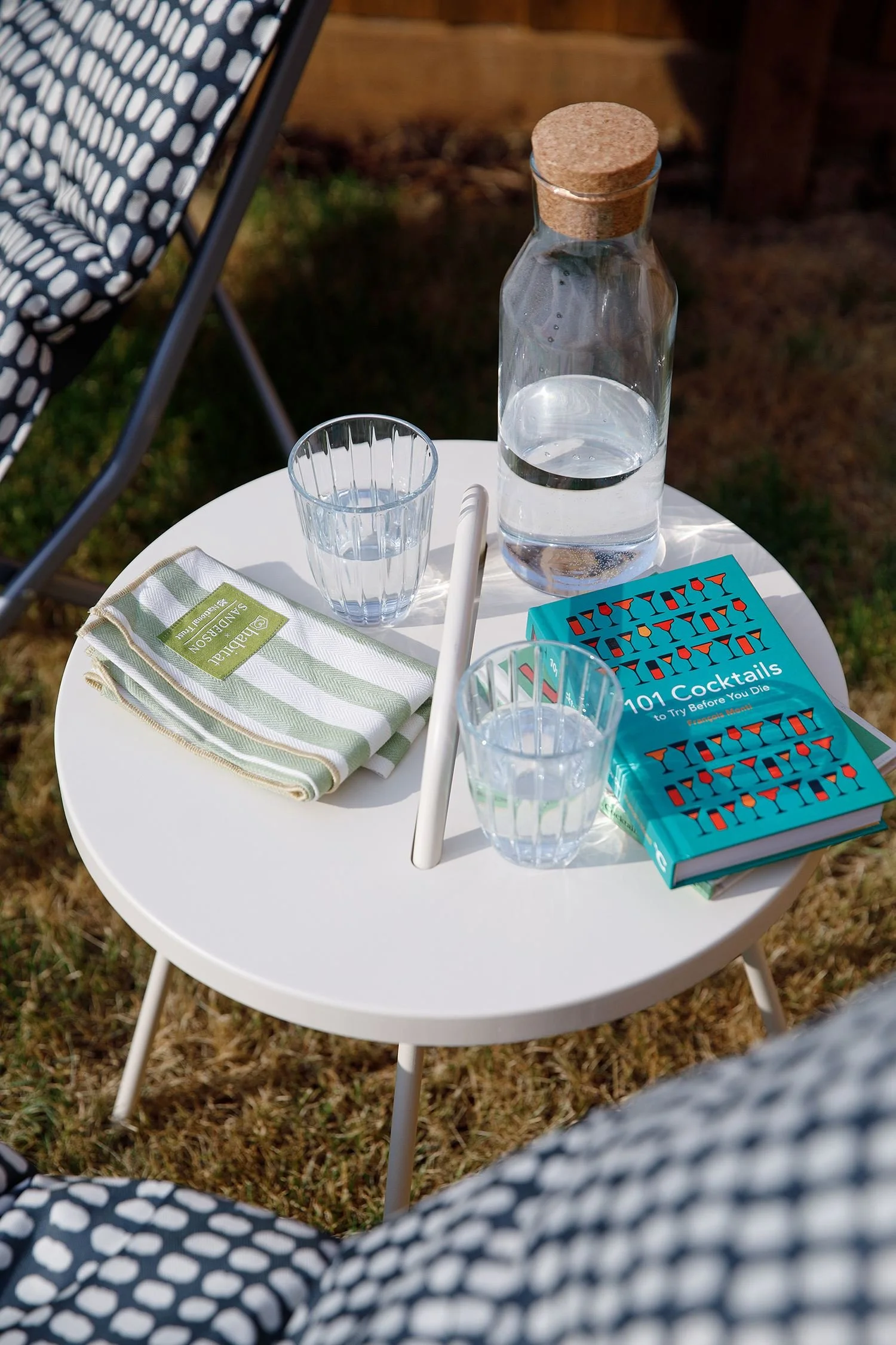 A small round white table outdoors with a water carafe, two glasses of water, a blue book titled 'Drinks & Cocktails,' a green and white checked cloth, and a white straw. In the background, there is a deck, some grass, and a part of a black and white