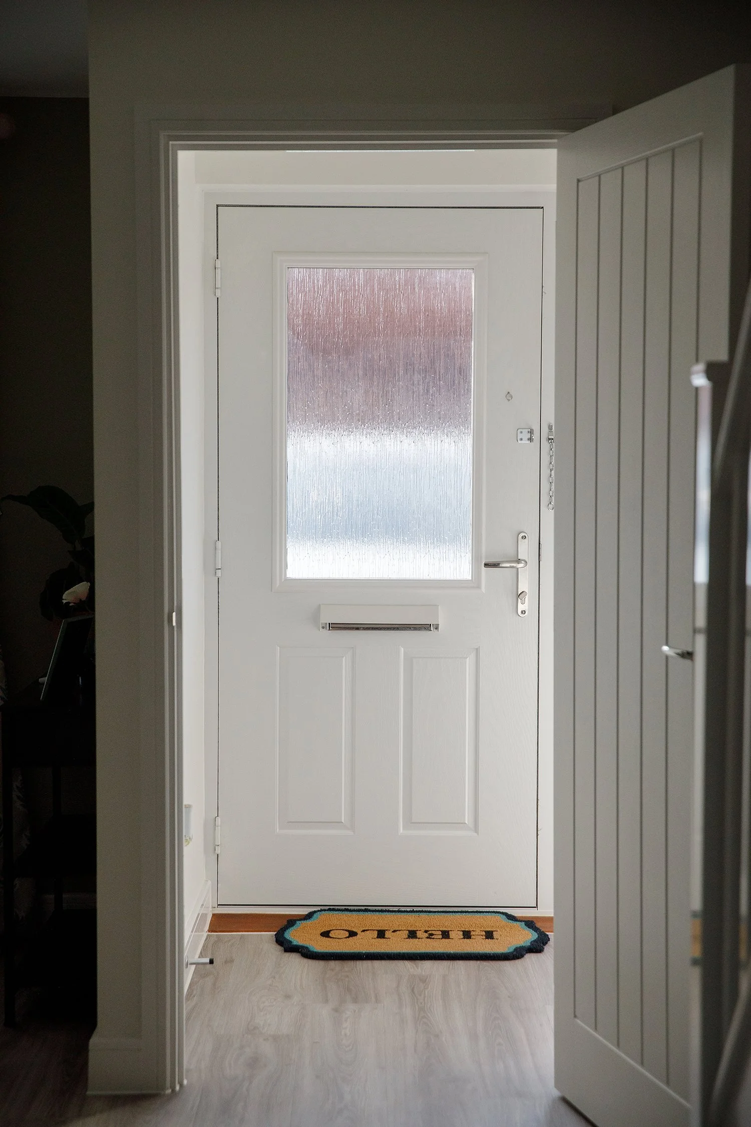 View through a doorway to a frosted glass front door with a mail slot, a lock, and a chain latch. There is a doormat with 'HELLO' in front of the door, and the floor is light-colored wood.