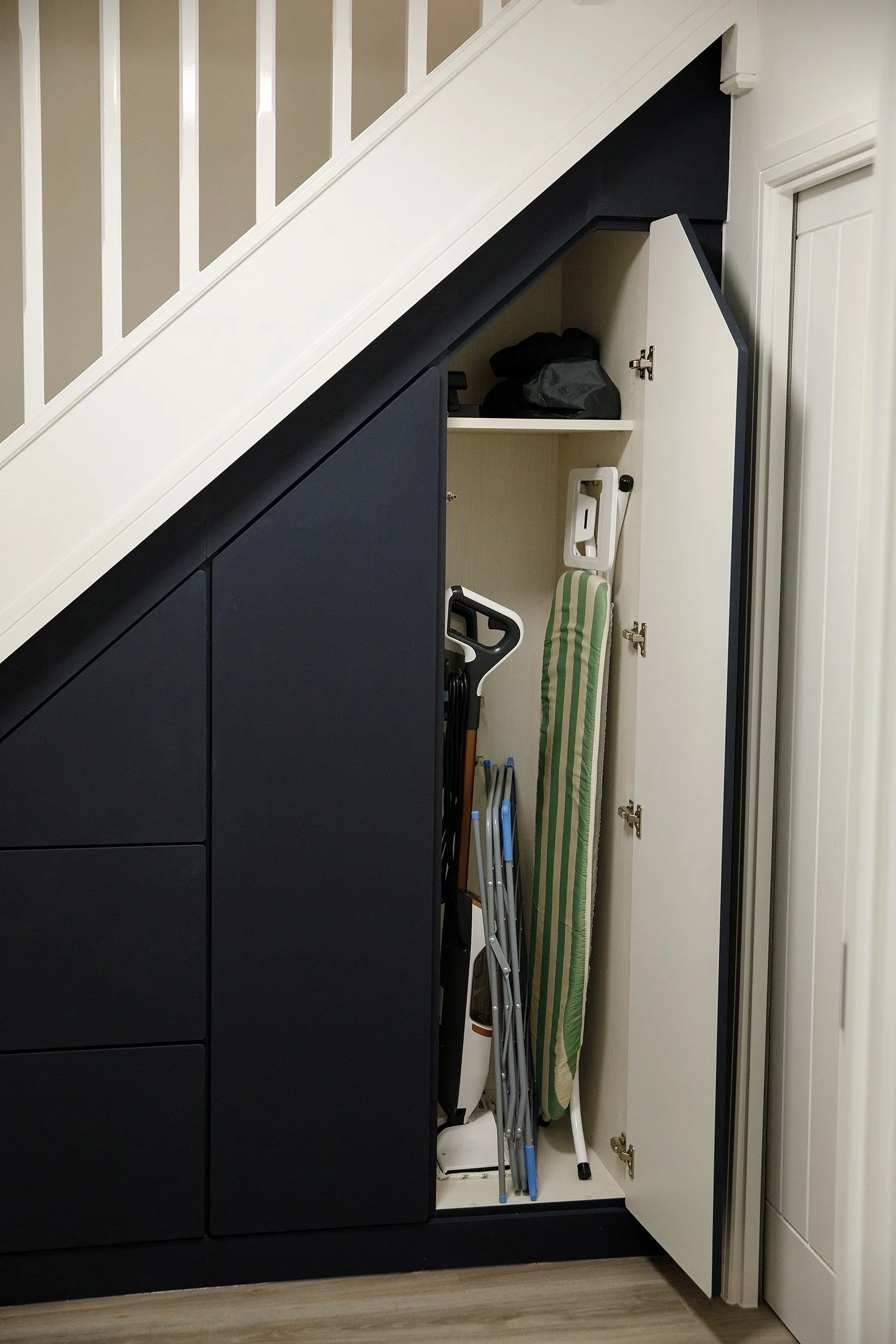 A closet under the staircase contains cleaning supplies, an ironing board, and foldable step ladders. The closet door is open, revealing stored items.