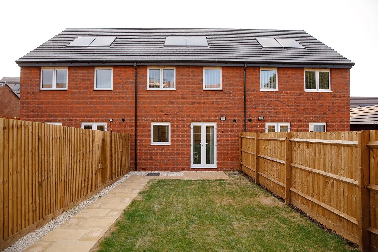 The rear view of a three-story brick house with a gray tiled roof and solar panels, a fenced backyard with a concrete and grass lawn, and back door with patio.