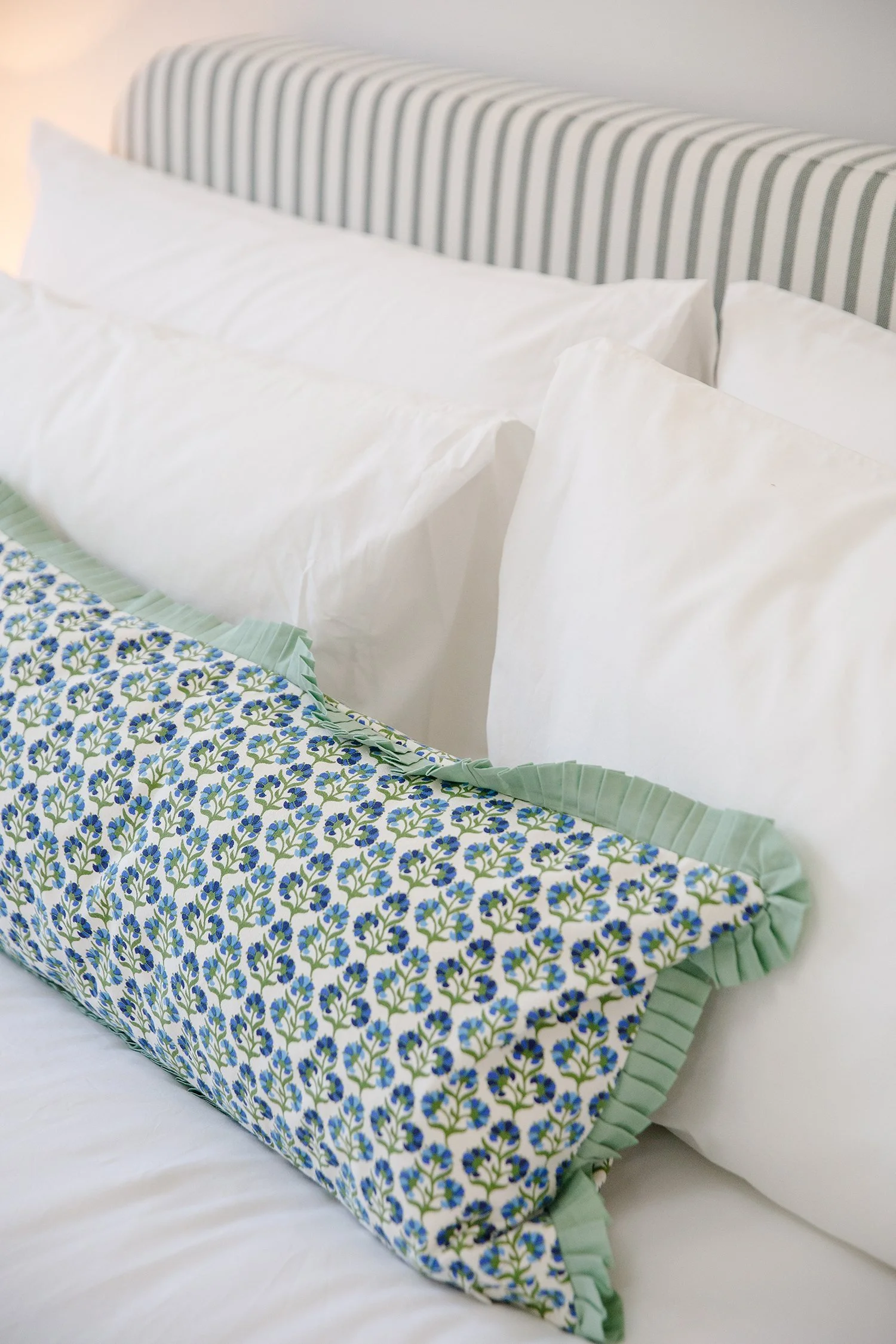 Decorative pillow with a floral pattern and pleated edges on a white bed, with white pillows and a striped headboard in the background.