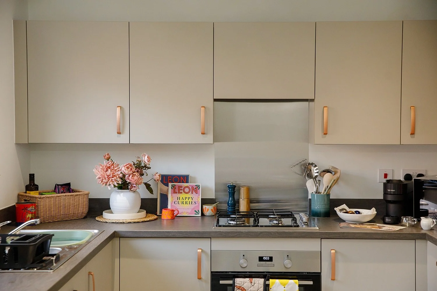 Kitchen with beige cabinets, black countertop, a stove, a white vase with pink flowers, books, and various kitchen items.